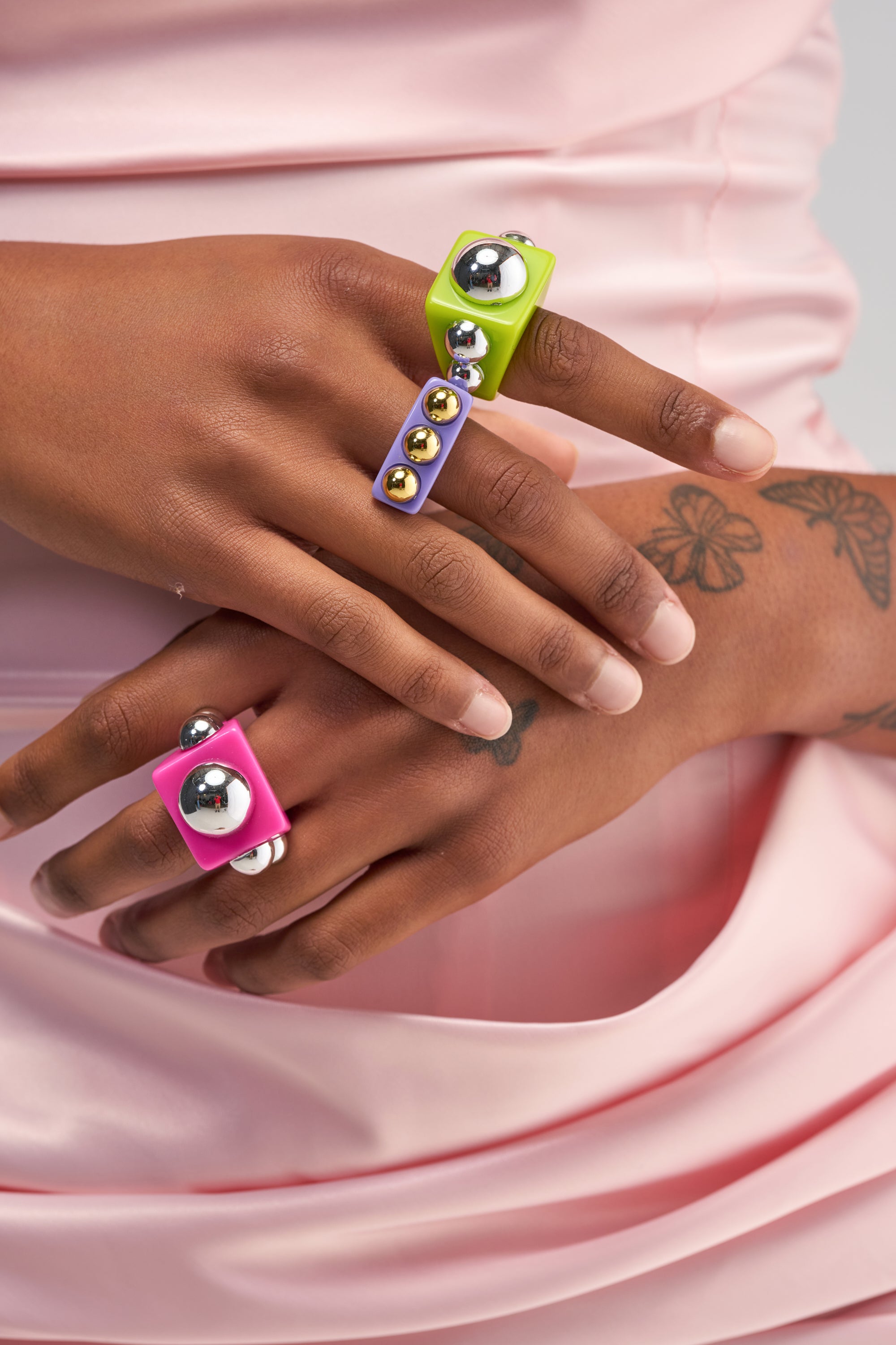 A close-up of tattooed hands adorned with the SPACE CANDY RING SET—colorful, chunky rings in pink, green, and purple with metallic accents. The person is wearing shiny light pink ruched fabric, highlighting these bold Y2K statement rings and maximalist jewelry for an eye-catching look.