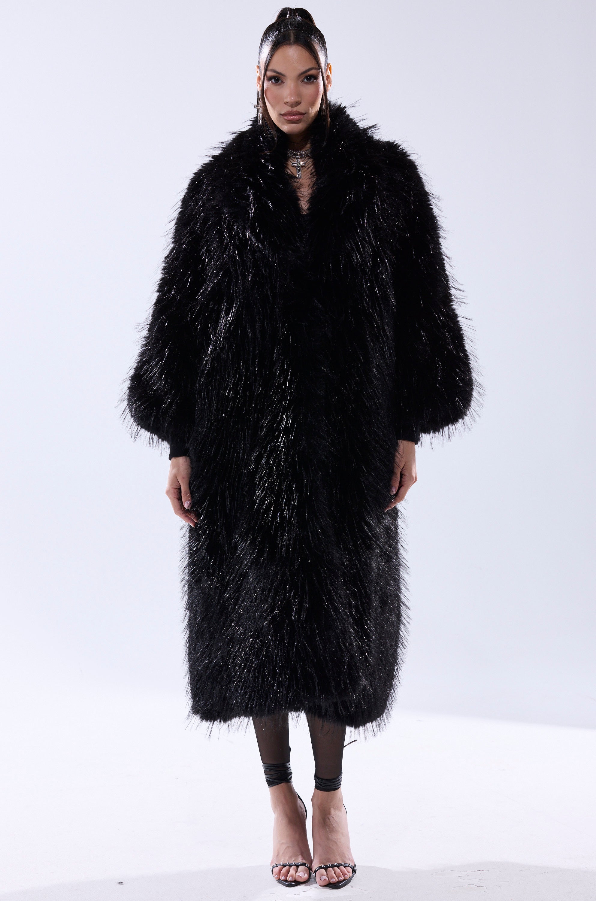 A woman exudes confidence in the TSUNAMI FAUX FUR COAT IN BLACK, styled over black leggings and open-toe heels. Her hair is straight and sleek as she poses against a plain background.