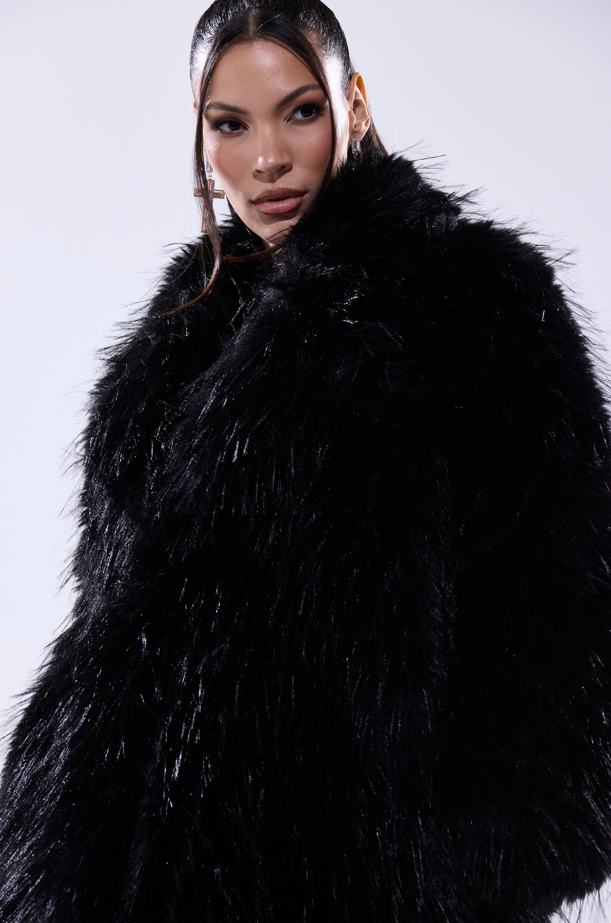 A woman with straight, dark hair and defined makeup models the TSUNAMI FAUX FUR COAT IN BLACK, posing confidently against a plain light background.