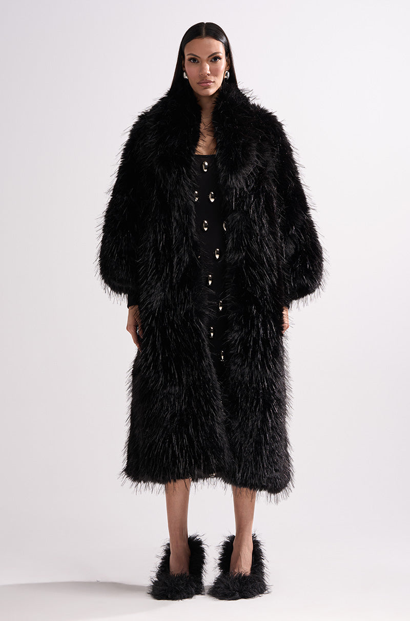 A woman stands facing forward in the TSUNAMI FAUX FUR COAT IN BLACK over a black dress with gold buttons and matching black furry shoes, set against a plain white background.