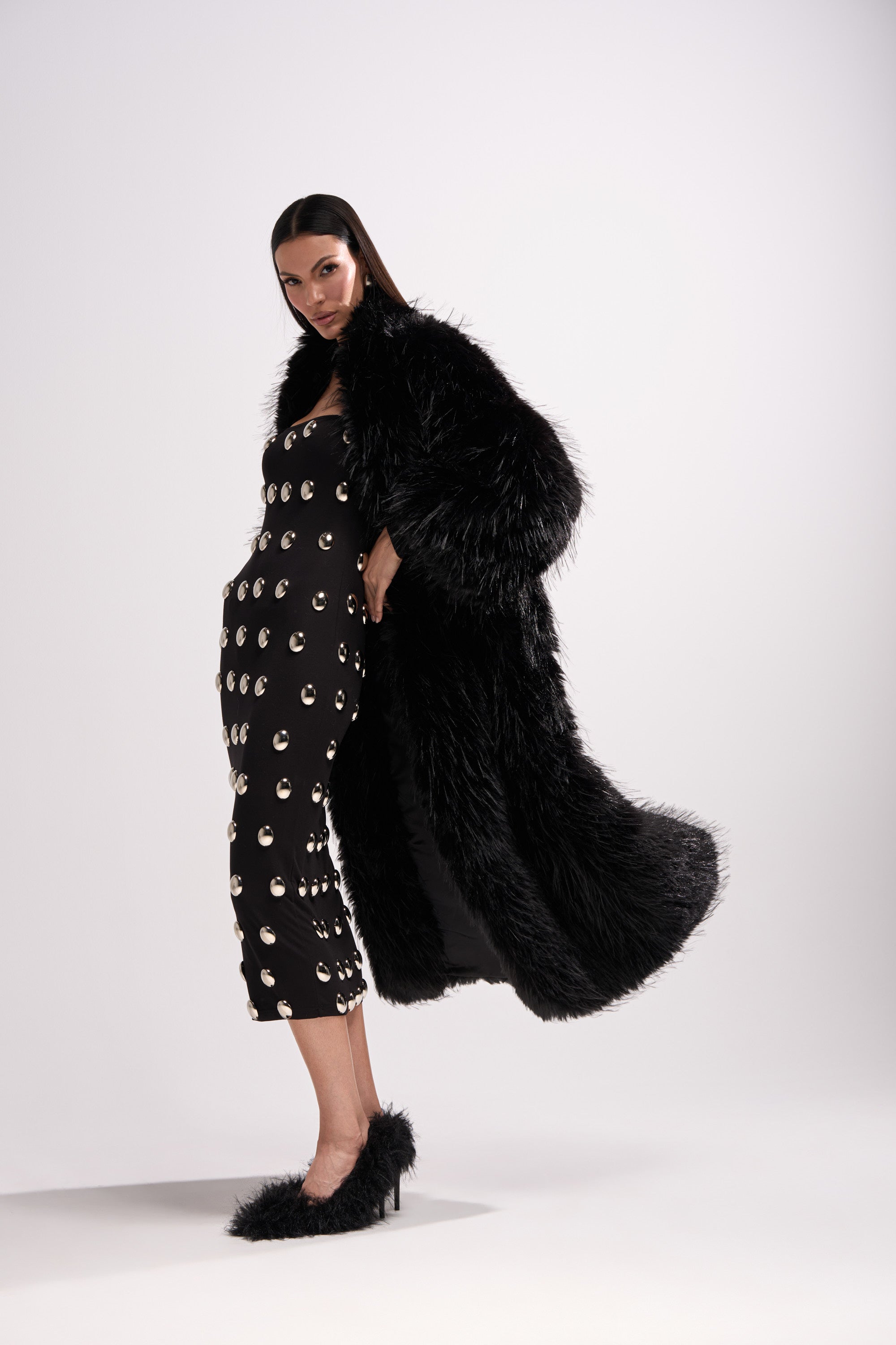 A woman models the TSUNAMI FAUX FUR COAT IN BLACK over a black dress with metallic eyelets and matching black furry heels, set against a plain white background.