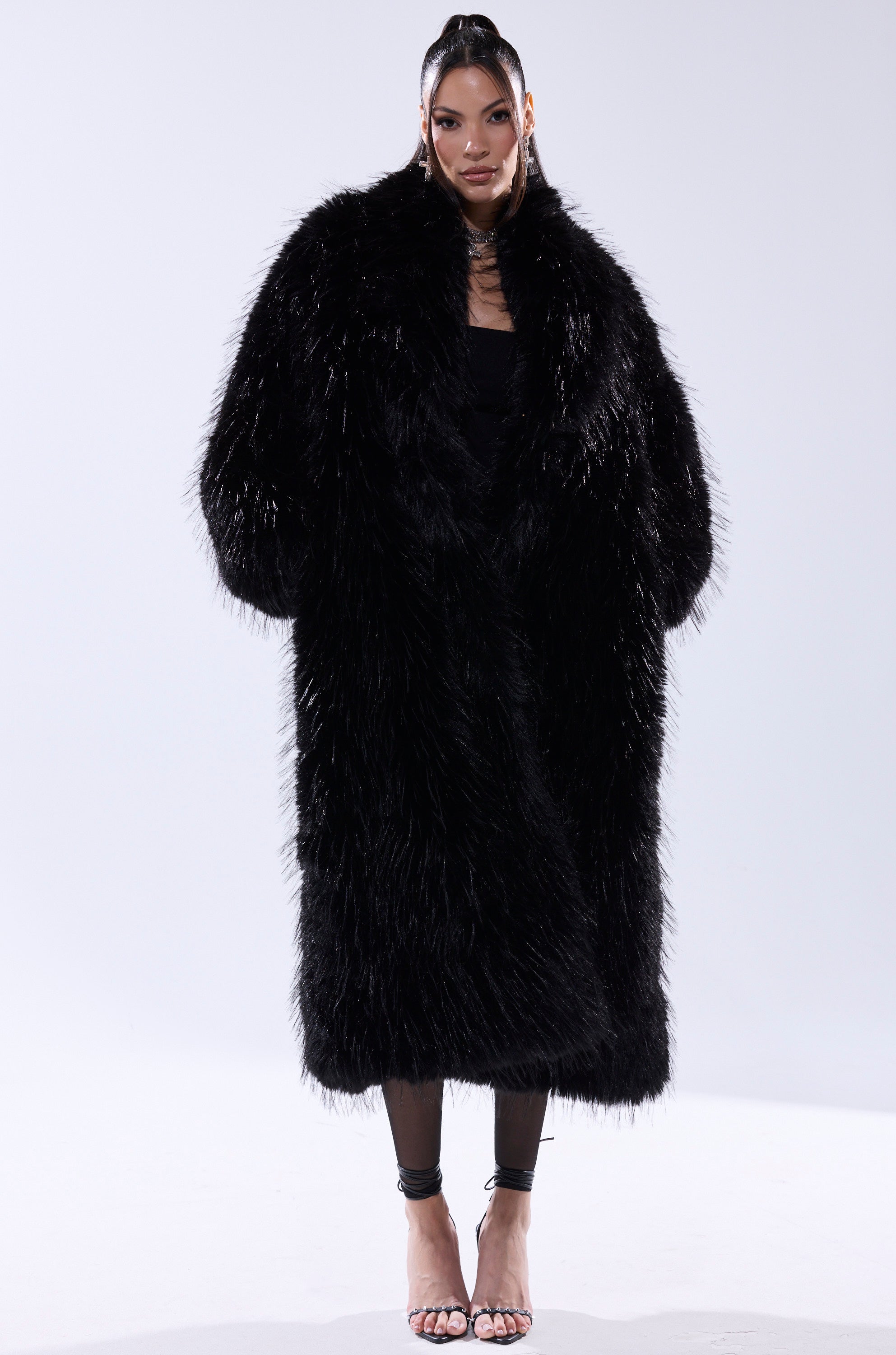 A woman faces forward wearing the TSUNAMI FAUX FUR COAT IN BLACK over black clothing and strappy high-heeled sandals, set against a plain white background.