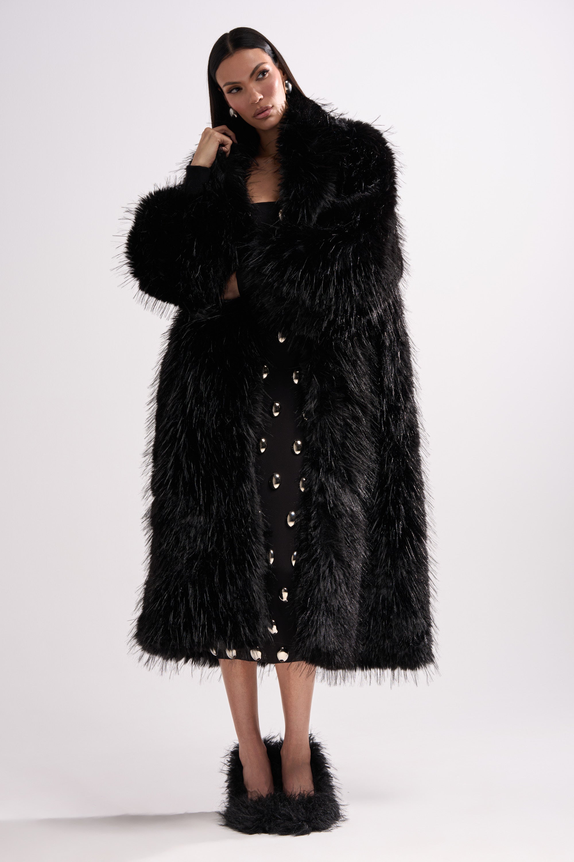A woman wearing the TSUNAMI FAUX FUR COAT IN BLACK over a black dress with metallic eyelets and matching fuzzy shoes, standing against a white background.