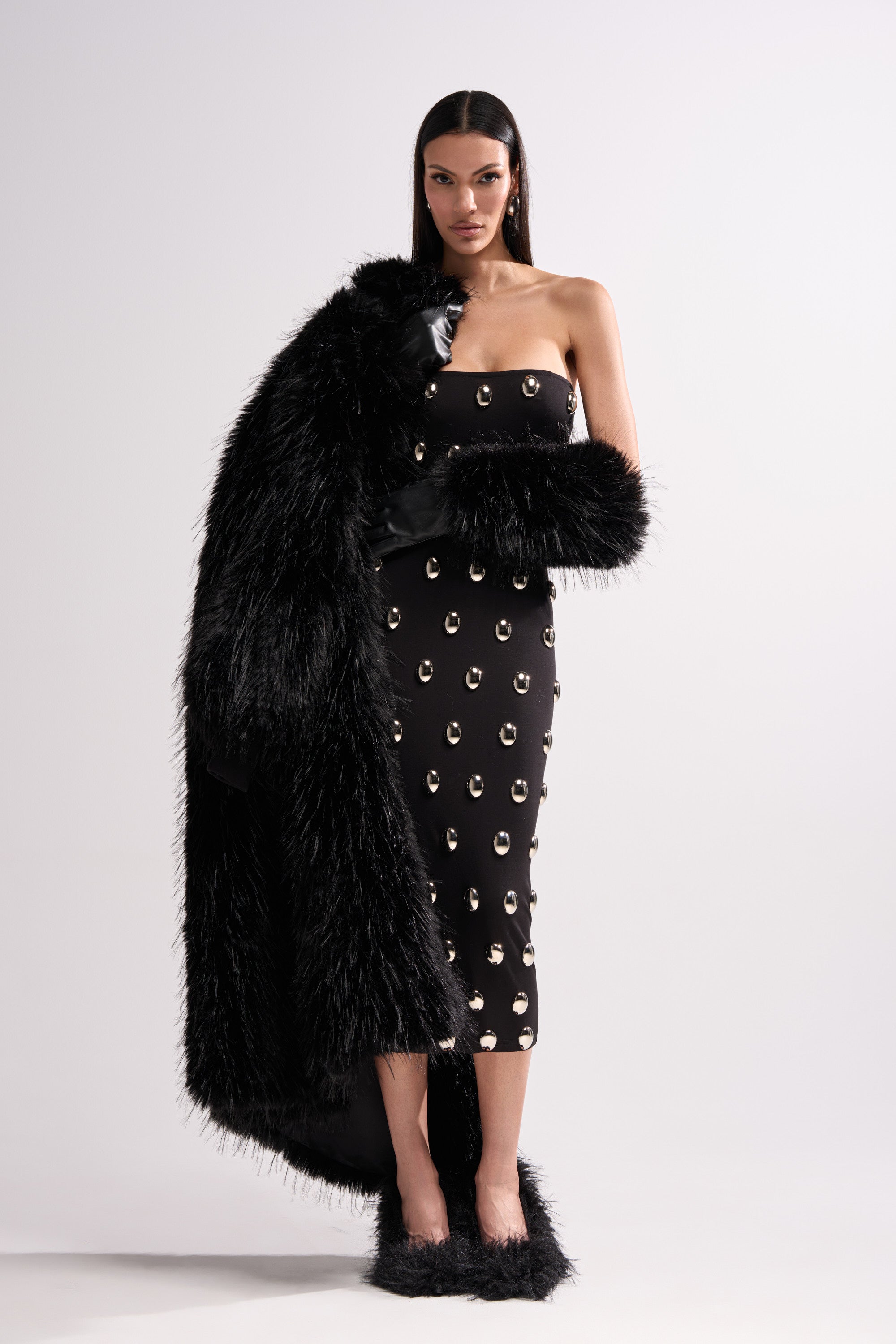 A woman poses against a plain white background, draping the TSUNAMI FAUX FUR COAT IN BLACK over one shoulder, paired with a strapless black dress with metallic accents and fluffy black shoes.