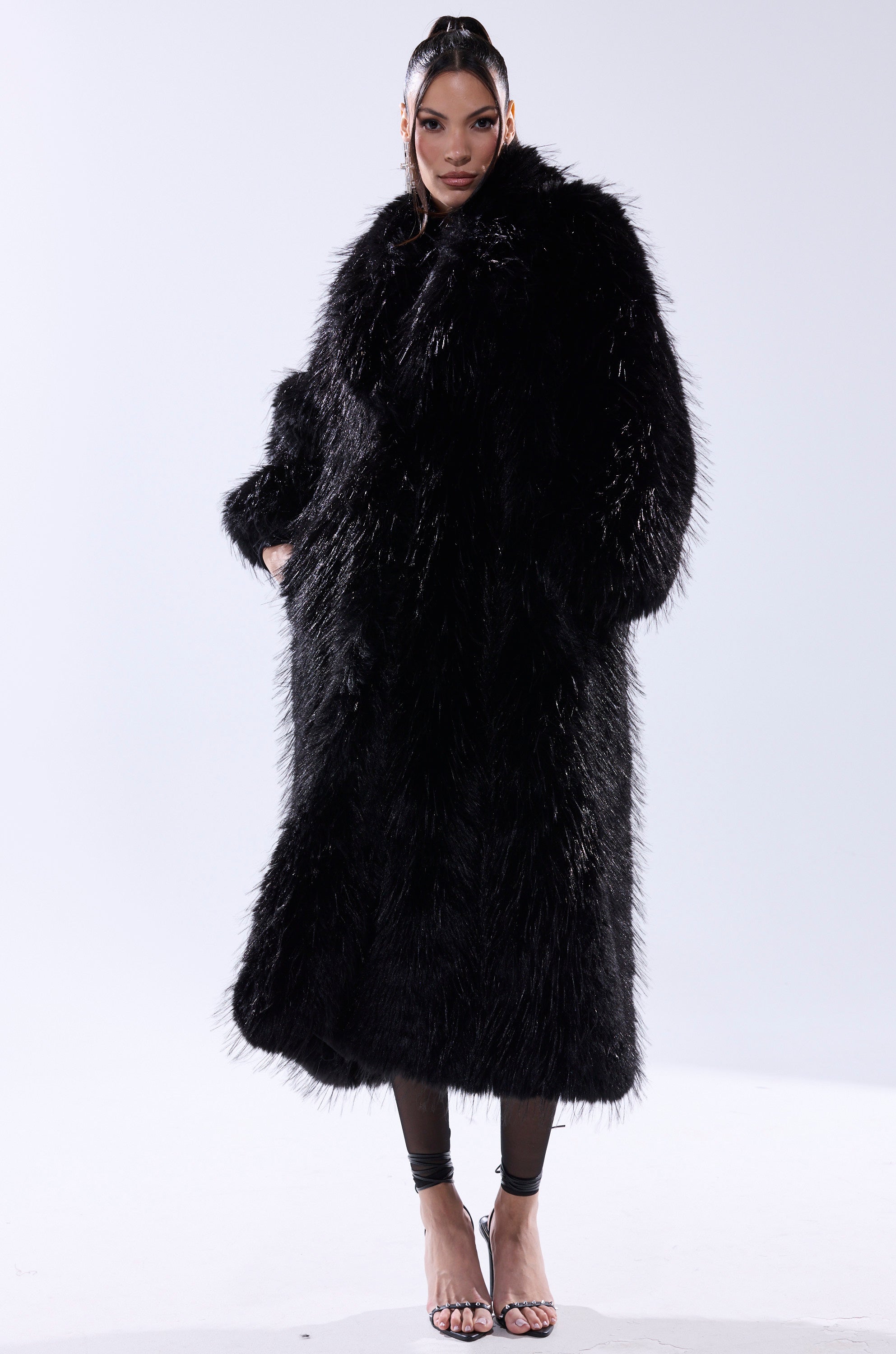 A woman stands against a plain background wearing the TSUNAMI FAUX FUR COAT IN BLACK with her hands in the pockets. She has sleek, styled hair and pairs the look with strappy high-heeled sandals.