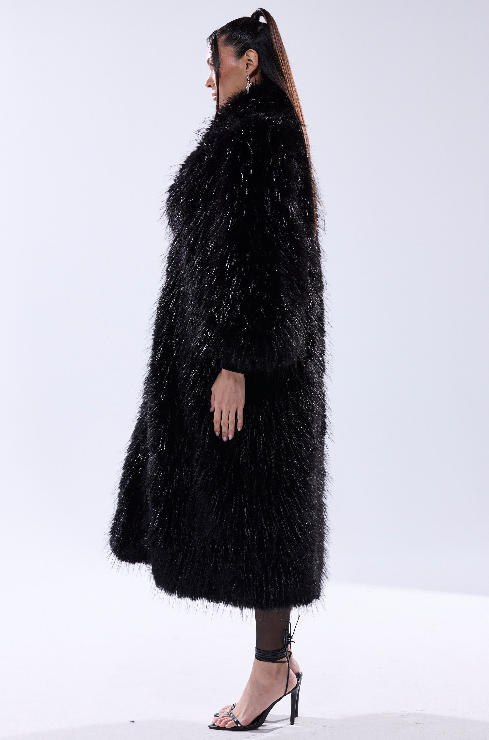 A person stands sideways against a plain white background, wearing the TSUNAMI FAUX FUR COAT IN BLACK paired with black high-heeled sandals.