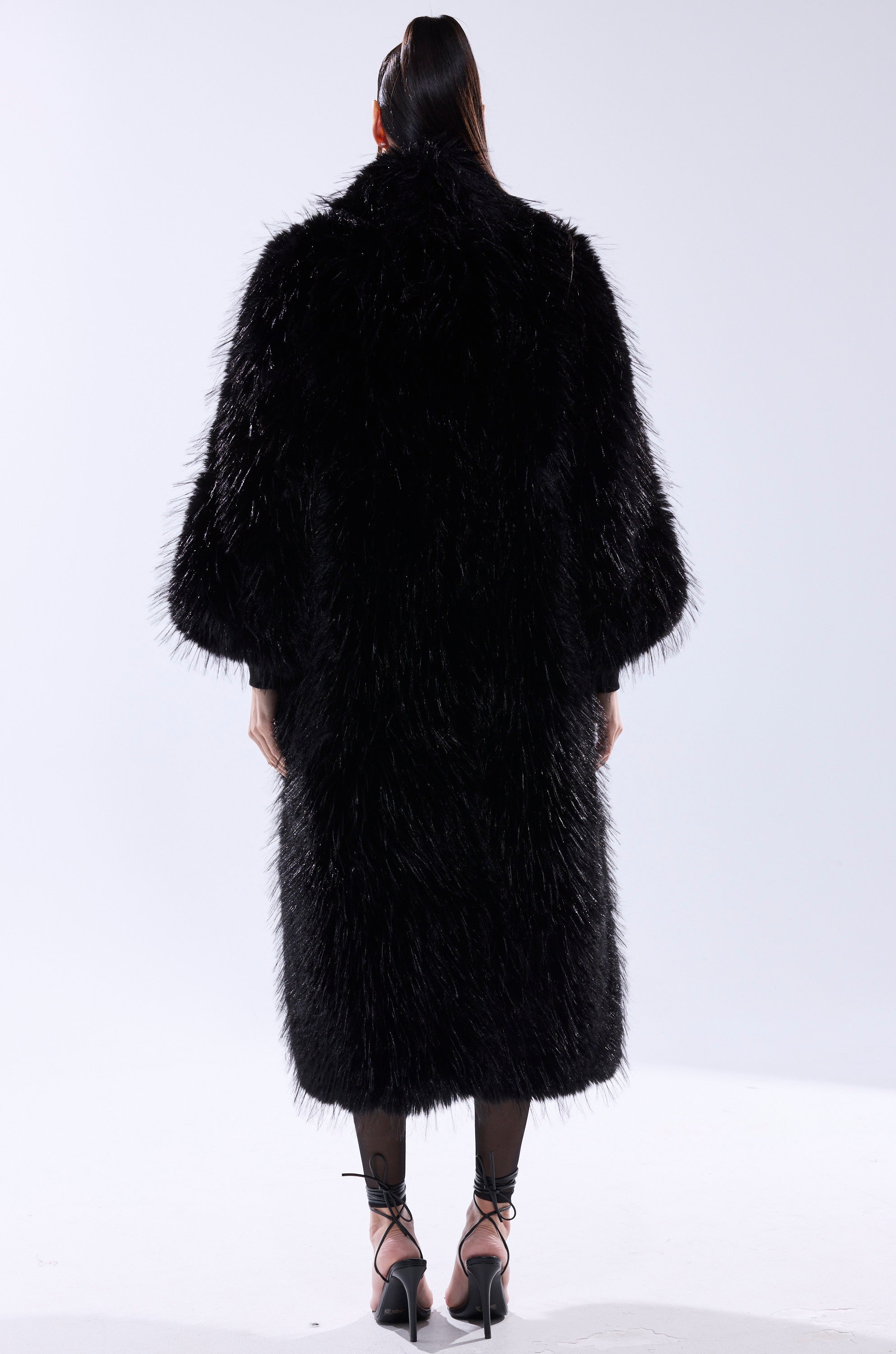 A person faces away, wearing the TSUNAMI FAUX FUR COAT IN BLACK with its voluminous texture, paired with black sheer tights and black strappy heels against a plain light background.