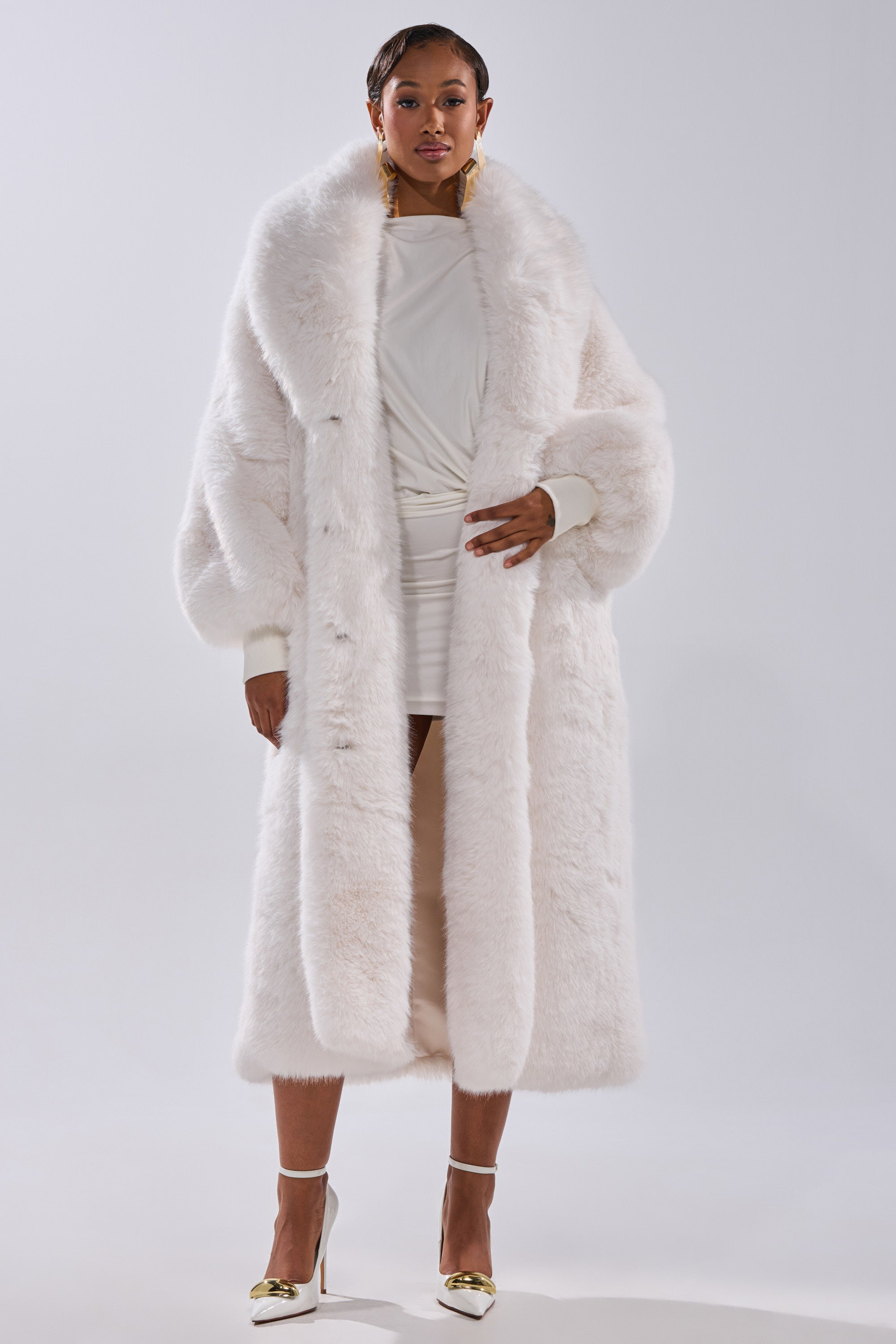 A woman exudes confidence in the TSUNAMI FAUX FUR COAT IN WHITE styled over a short white dress, complemented by gold earrings and white high heels with gold accents, set against a simple light background.