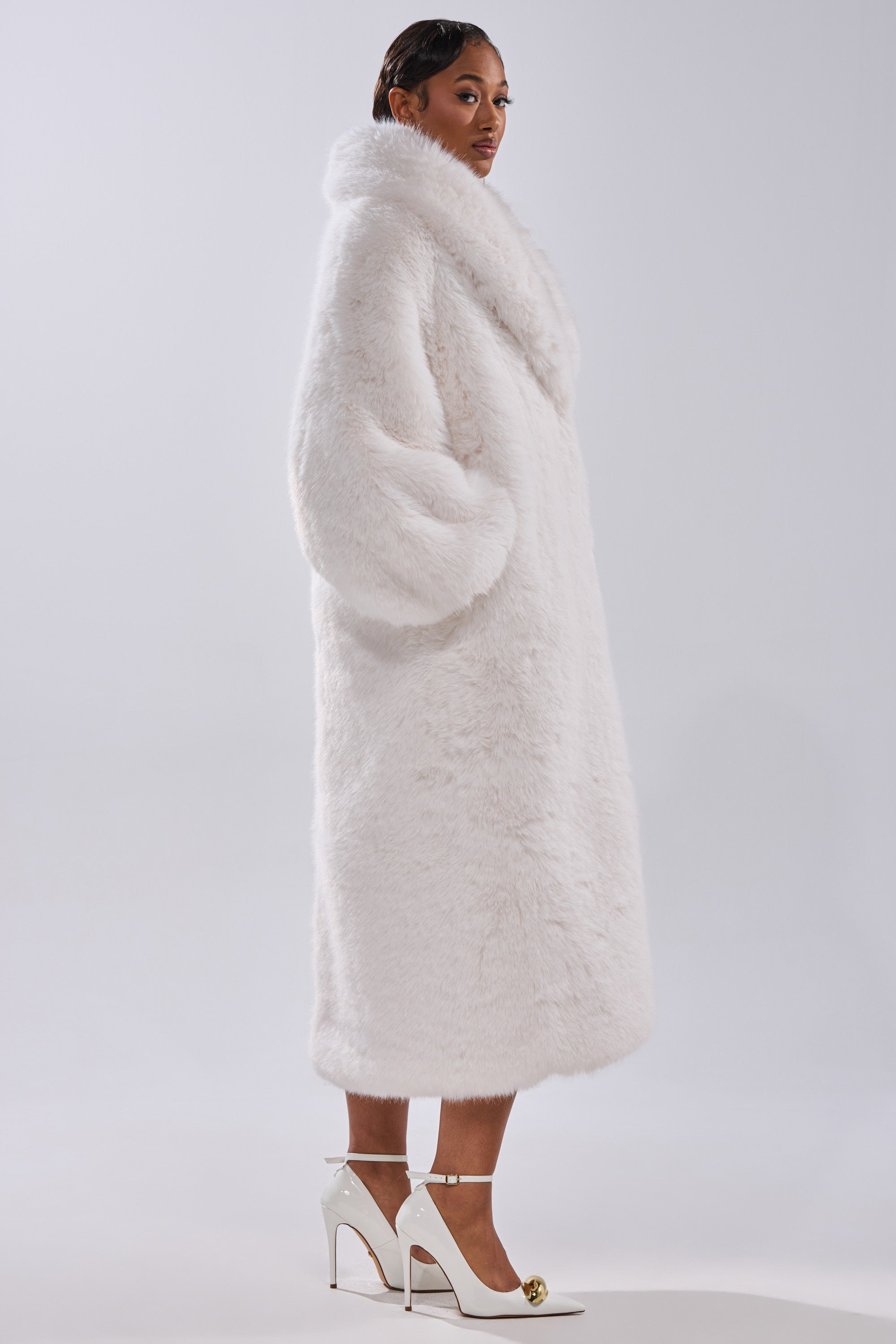 A woman stands in profile against a plain light background, wearing the TSUNAMI FAUX FUR COAT IN WHITE styled as a longline silhouette, paired with white high-heeled shoes featuring gold accents.