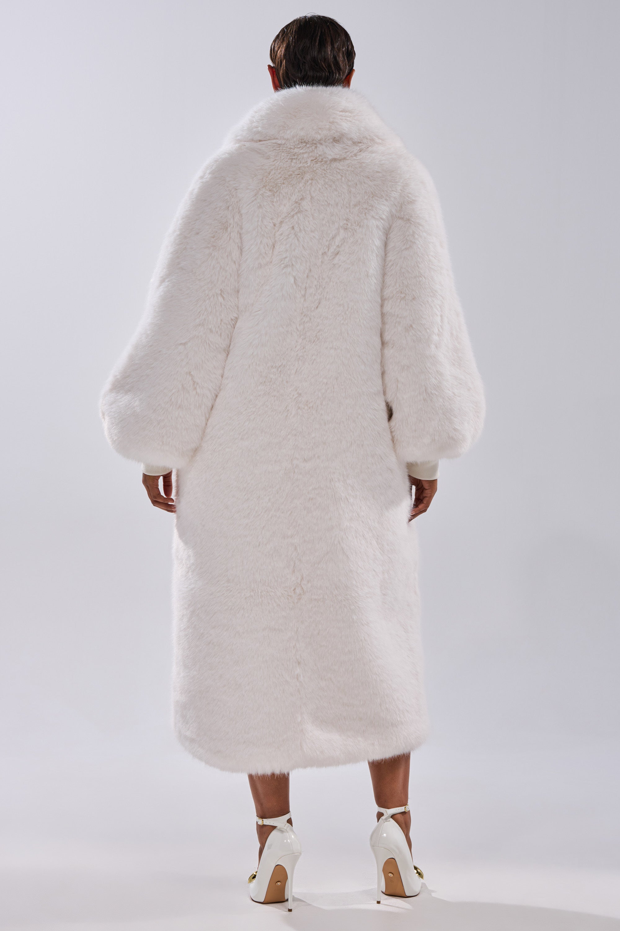 A person with short dark hair stands with their back to the camera, wearing the TSUNAMI FAUX FUR COAT IN WHITE, featuring a longline silhouette, wide sleeves, and high collar, paired with white ankle-strap high heels on a plain light background.