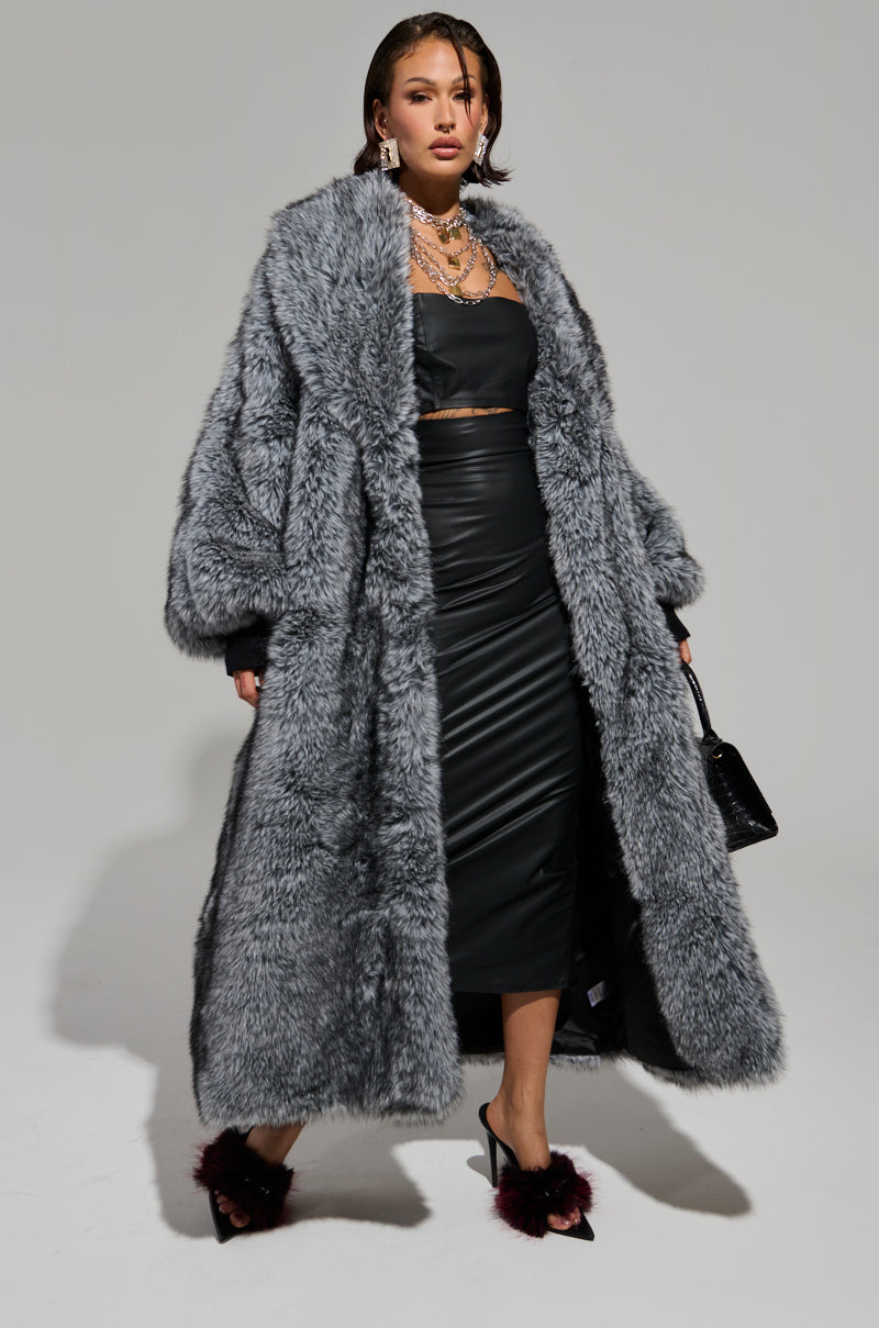 Dressed with confidence, a person showcases the TSUNAMI FAUX FUR TRENCH in gray, layered over a sleek black outfit. Their look is elevated with large earrings, a layered necklace, and feathered high heels. A black purse is elegantly positioned at their side. 1