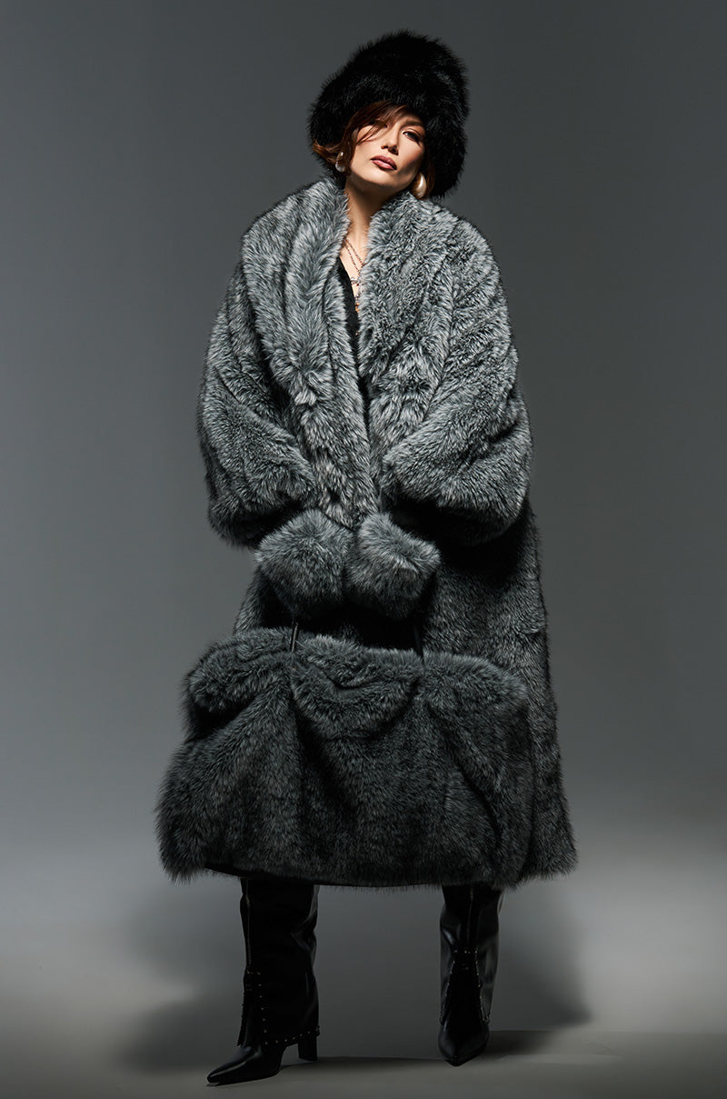 A person poses confidently in the oversized TSUNAMI FAUX FUR TRENCH, a luxurious gray faux fur coat paired with a matching black fur hat. Their sleek black knee-high boots enhance the ensemble, while the plain gray background highlights their stylish winter attire. 0