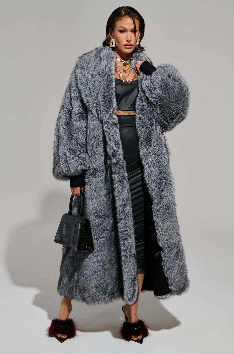 A person is wearing the TSUNAMI FAUX FUR TRENCH over a black satin dress. They hold a black handbag and wear high heels with fur details. Their hair is styled back, adorned with multiple necklaces, and the stylish coat perfectly complements the plain background. 4