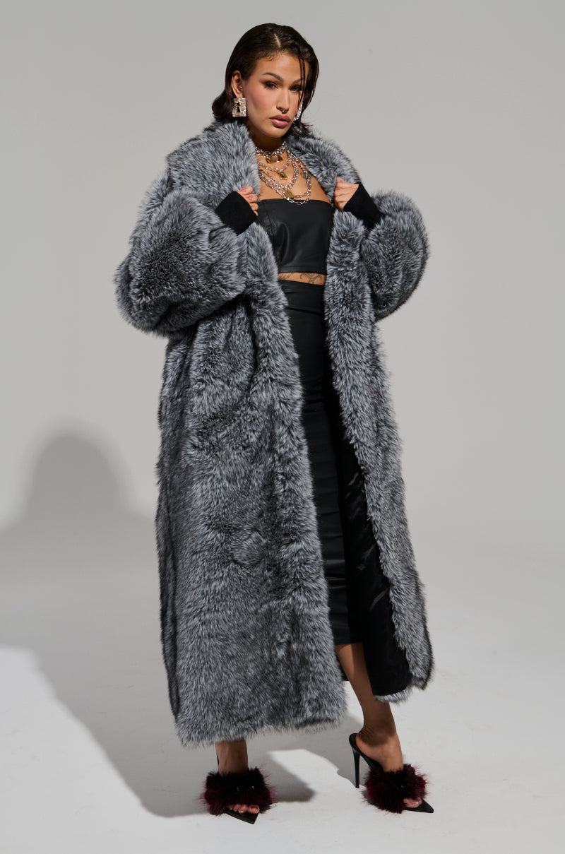 Against a plain background, a person confidently wears the TSUNAMI FAUX FUR TRENCH over a sleek black outfit. They complement the look with elegant jewelry and feathered heels, projecting a bold and stylish vibe. 5