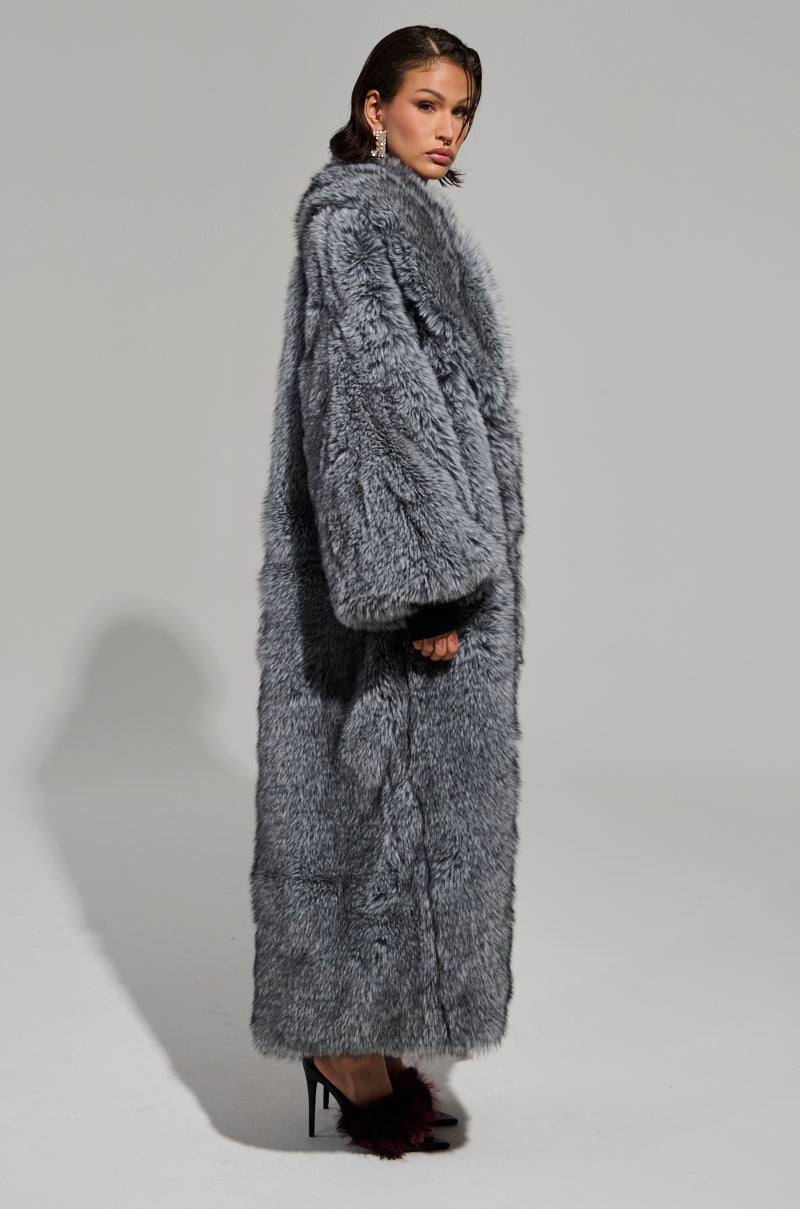 A person stands against a plain background, elegantly draped in the TSUNAMI FAUX FUR TRENCH with an oversized fit. The long, fluffy gray coat beautifully complements their black high-heeled shoes. They gaze to the side, with medium-length hair softly framing their face and hands partially tucked into the trench's pockets. 6