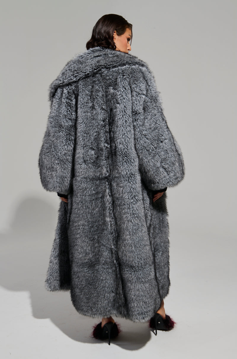 A person is seen from the back wearing the TSUNAMI FAUX FUR TRENCH, which features an oversized gray design with a large collar. They stand against a plain, light-colored background, also wearing black high-heeled shoes adorned with fur details. 7