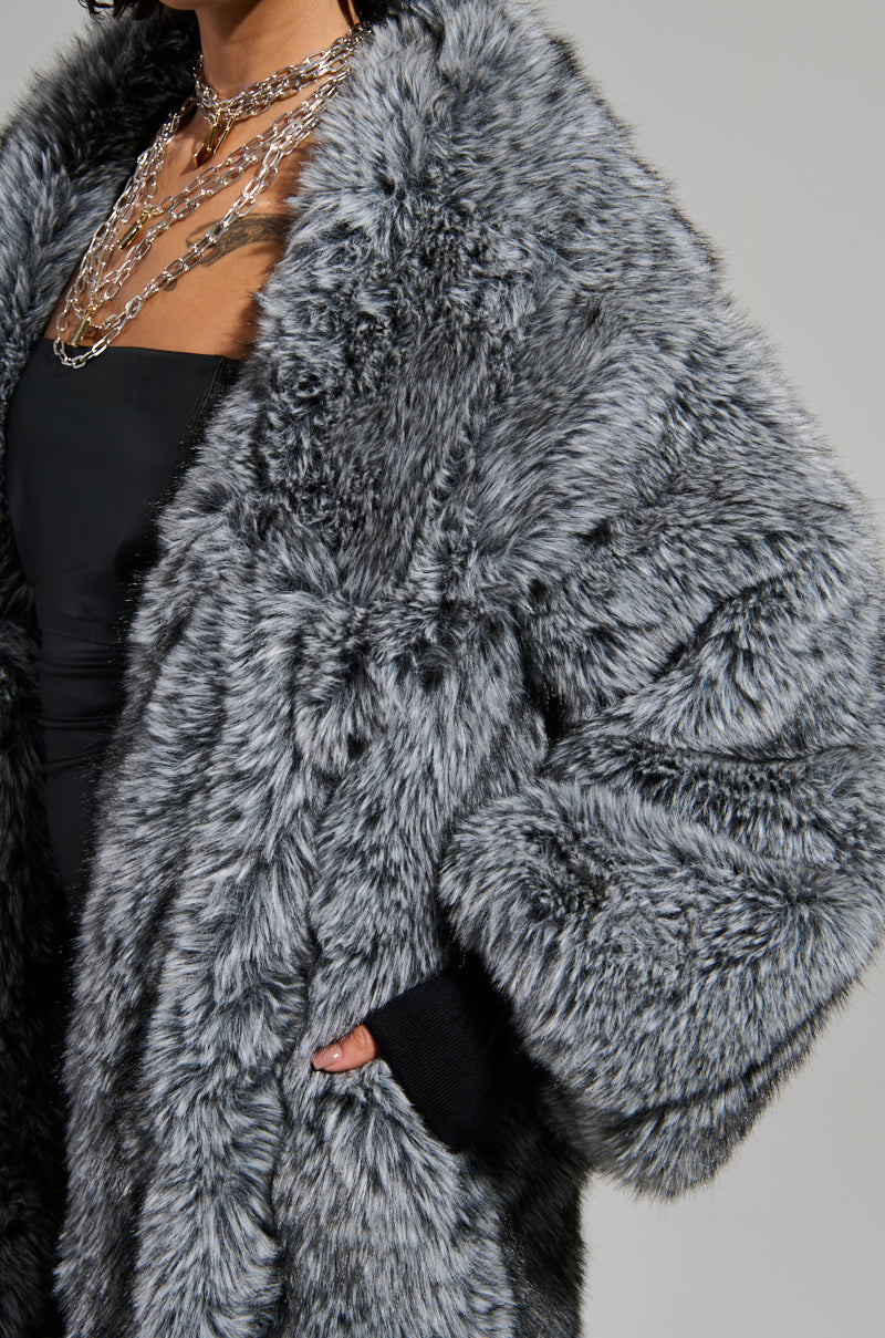 Dressed in the opulent TSUNAMI FAUX FUR TRENCH coat with an oversized fit, a person stylishly places their hand in the pocket. They pair this luxurious piece with layered silver chain necklaces over a black top, creating a harmonious and chic winter look. 8