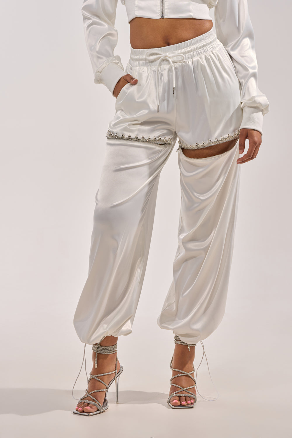 Wearing the CALL BACK CUTOUT SATIN JOGGER in shiny white with decorative trim and knee cutouts, paired with a matching cropped jacket and strappy heels, stands against a plain light background. 0