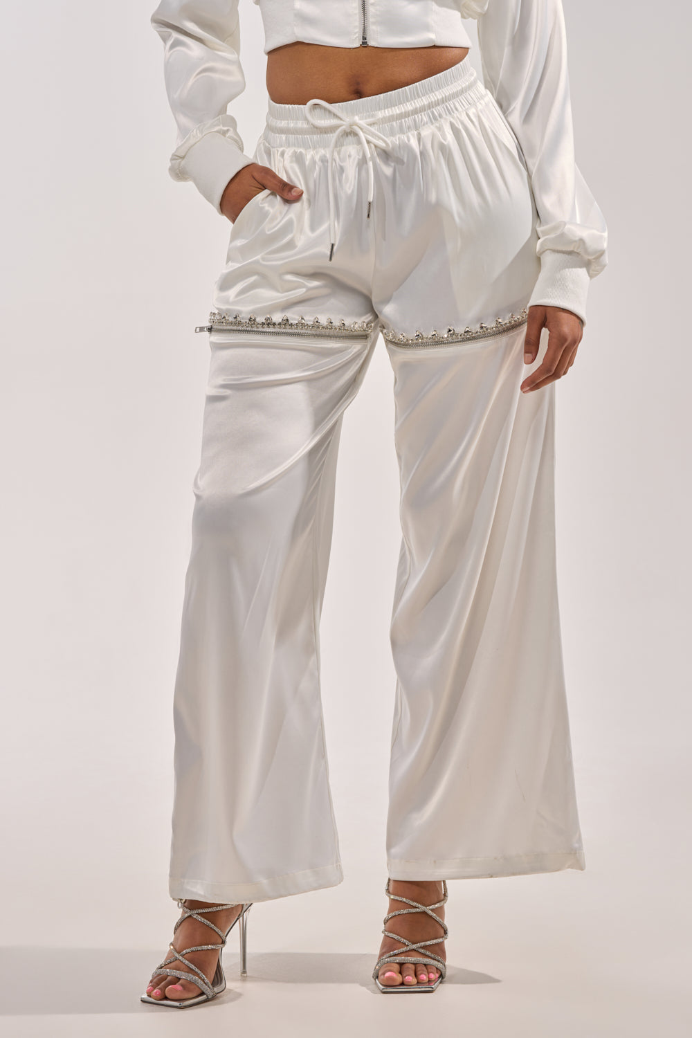 Someone in the CALL BACK CUTOUT SATIN JOGGER—loose-fitting white satin joggers with a drawstring waist and pearl details—paired with a matching cropped jacket and silver strappy high-heeled sandals. 1