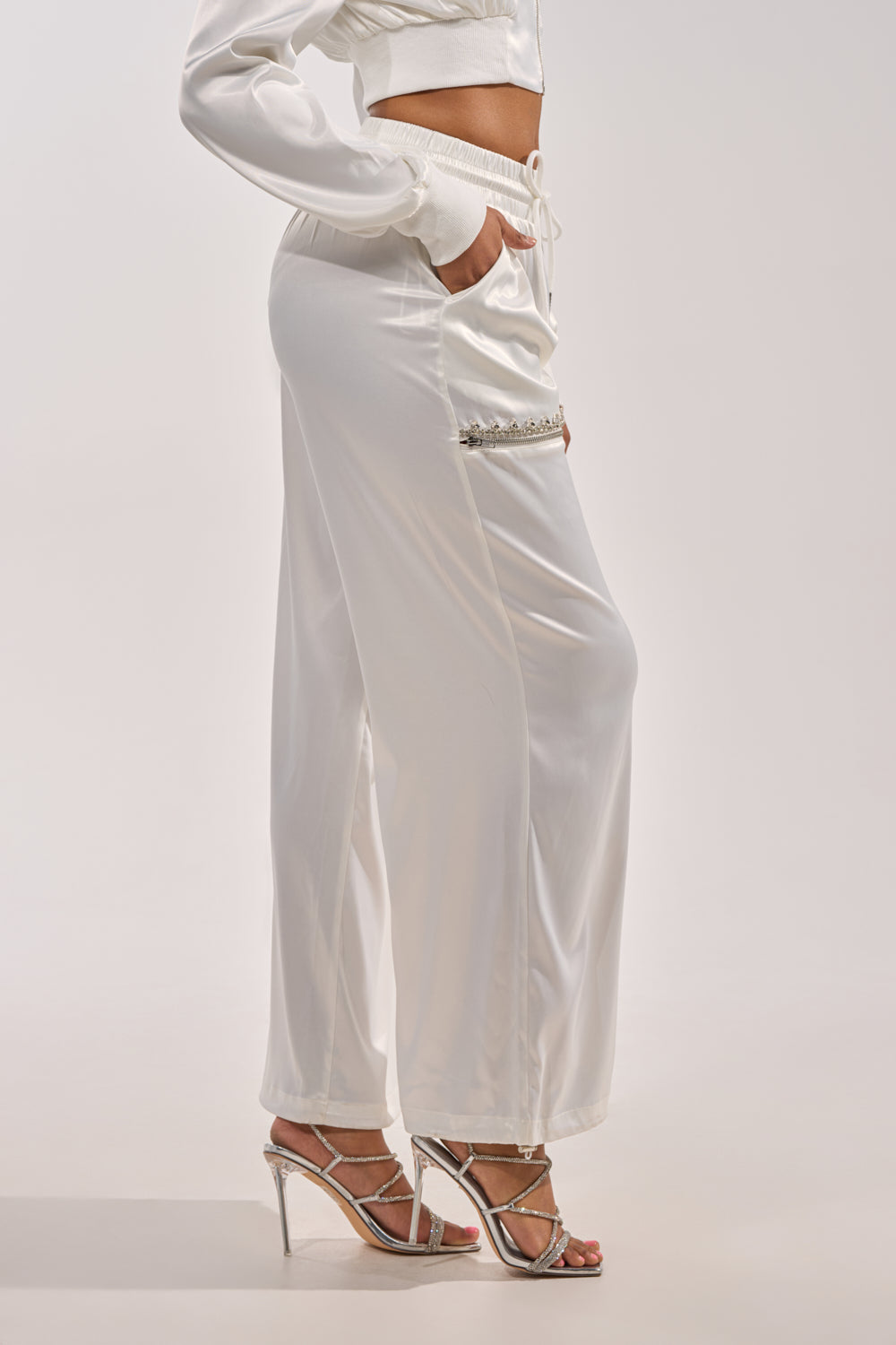 Wearing the CALL BACK CUTOUT SATIN JOGGER with a drawstring waist and matching cropped jacket, this person stands sideways in silver high-heeled sandals, one hand in their pocket. 2