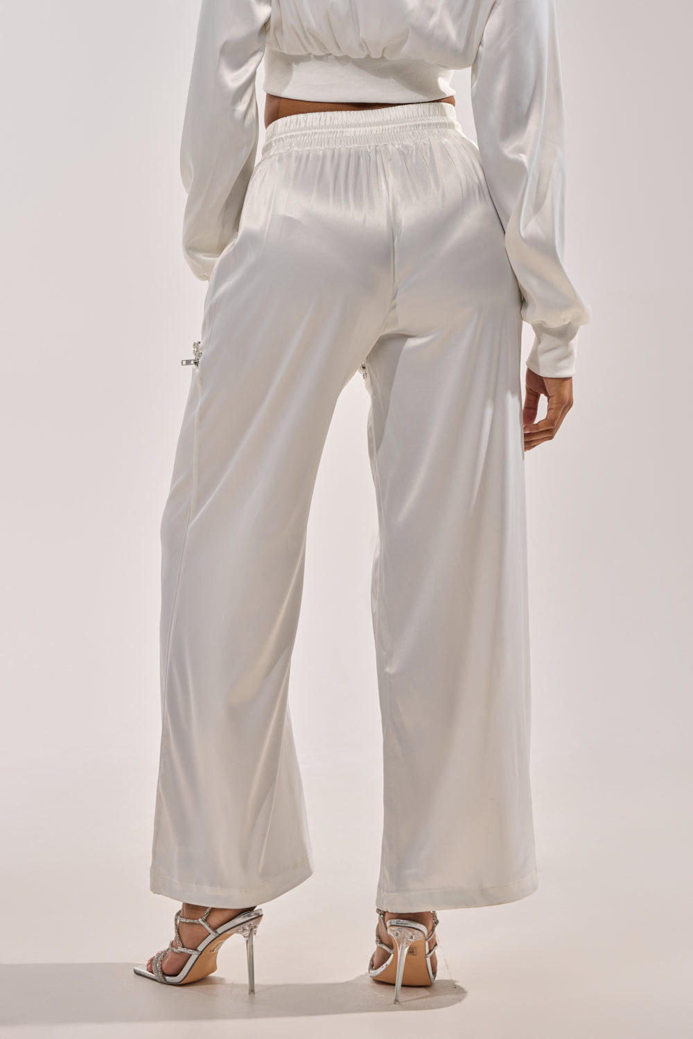 A person is seen from the back wearing CALL BACK CUTOUT SATIN JOGGER pants and silver high-heeled sandals against a plain light background. 3