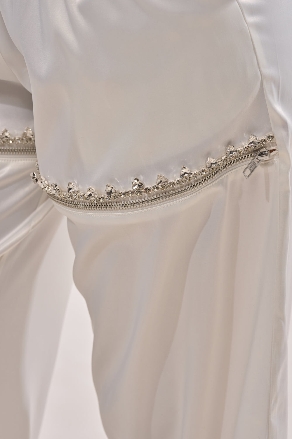 Close-up of the CALL BACK CUTOUT SATIN JOGGER, showcasing shiny satin fabric and a horizontal silver zipper with decorative beads or studs above it, emphasizing the detailed texture and high-waisted design. 6