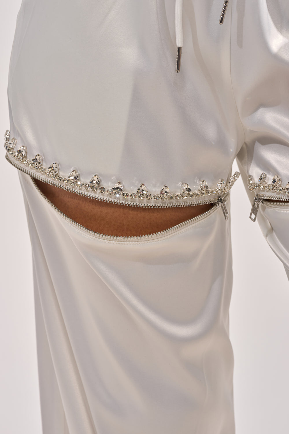 Close-up of the CALL BACK CUTOUT SATIN JOGGER featuring shiny white satin and a partially open decorative zipper trimmed with small pearl-like beads, adding an elegant touch and revealing a hint of skin underneath. 7