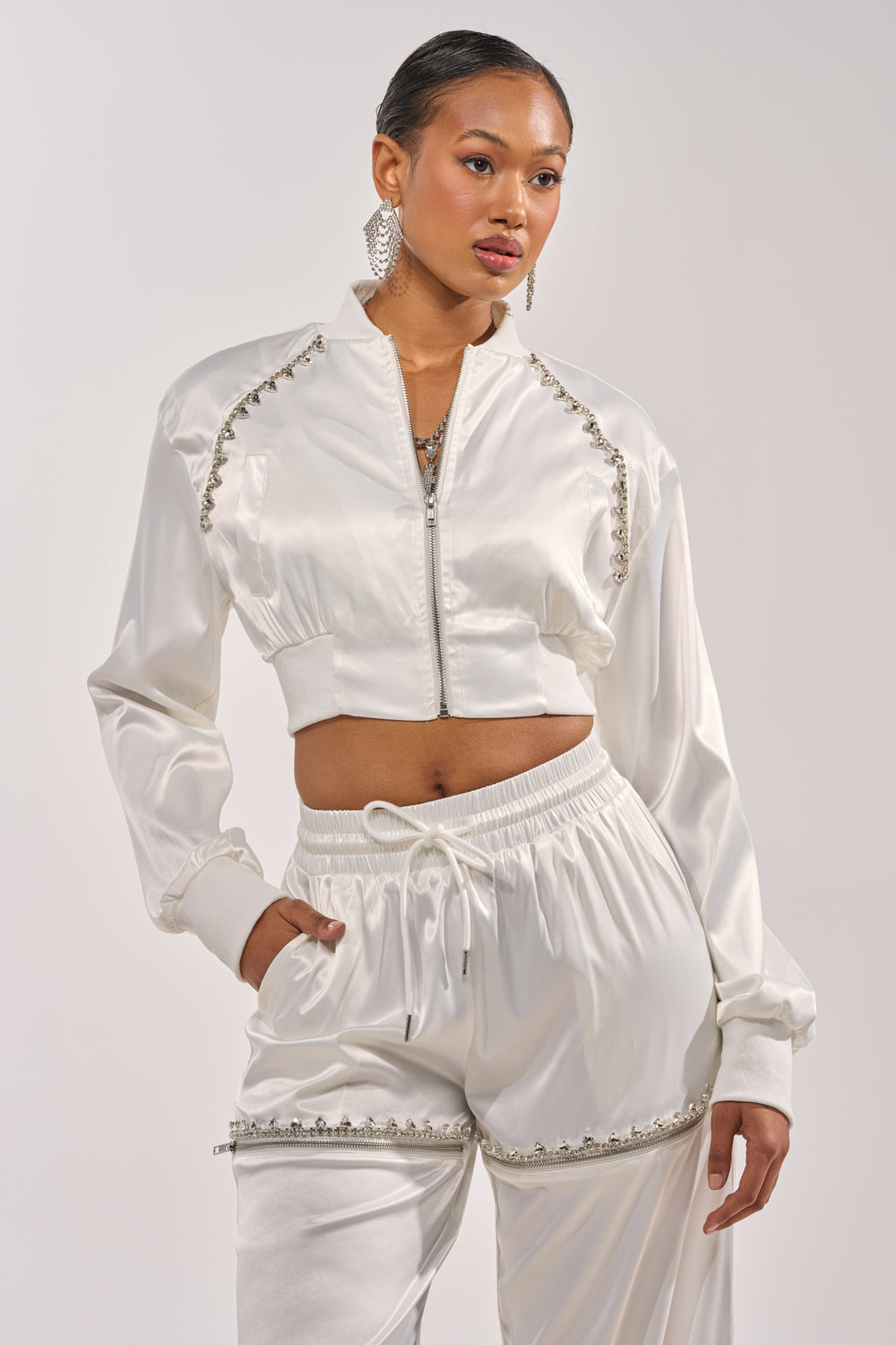 A woman models the CALL BACK SATIN BOMBER, a shiny white cropped jacket with jeweled details, paired with matching pants. She poses with one hand in her pocket against a plain light background. 0