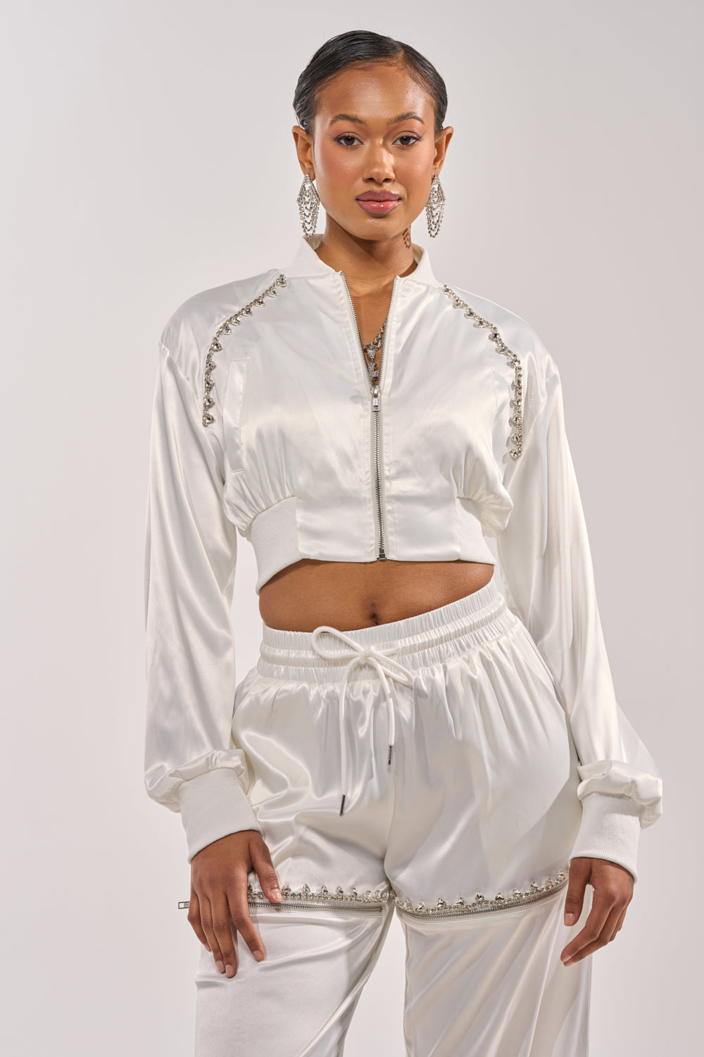 A woman with slicked-back hair and statement earrings poses confidently in the CALL BACK SATIN BOMBER— a white cropped jacket with silver embellishments— paired with matching joggers, set against a plain background. 3