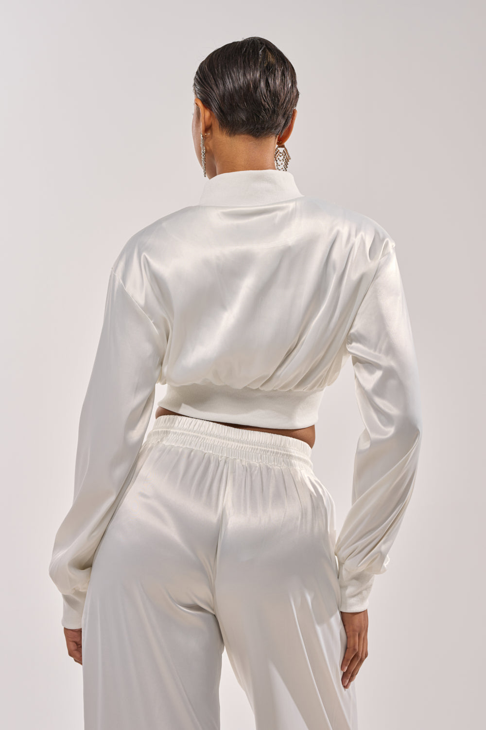 A person with short, slicked-back hair is seen from behind against a plain light background, wearing the CALL BACK SATIN BOMBER in white with matching high-waisted pants. 5
