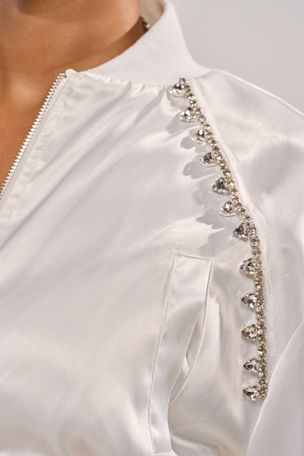 Close-up of the CALL BACK SATIN BOMBER, a white satin bomber jacket featuring a zipper and silver rhinestone embellishments along the shoulder seam. 8