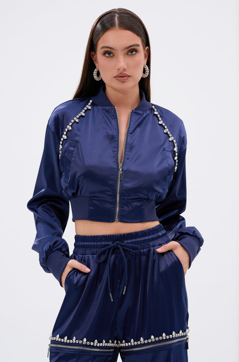 A woman stands confidently in the CALL BACK SATIN BOMBER—a shiny navy blue cropped satin jacket with pearl embellishments—paired with matching drawstring pants. She wears hoop earrings, has long straight hair, and poses hands-in-pockets against a white background.