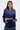A woman stands confidently in the CALL BACK SATIN BOMBER—a shiny navy blue cropped satin jacket with pearl embellishments—paired with matching drawstring pants. She wears hoop earrings, has long straight hair, and poses hands-in-pockets against a white background.