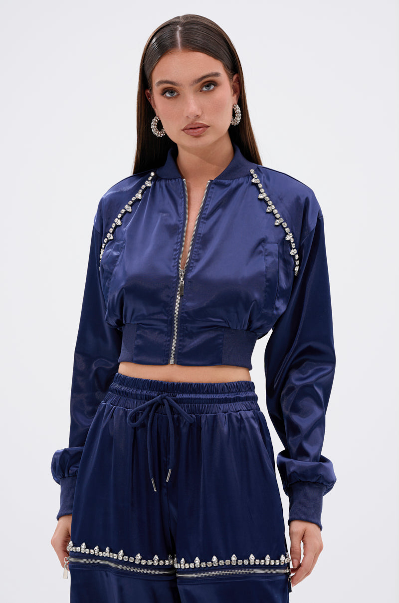 A woman wears the CALL BACK SATIN BOMBER, a shiny navy blue cropped jacket with pearl and rhinestone trim, paired with matching high-waisted pants. She has long straight hair, statement earrings, and a neutral expression.