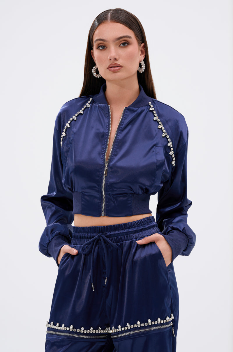 A woman wears the CALL BACK SATIN BOMBER, a shiny navy blue cropped satin jacket with pearl details and matching high-waisted pants. She has long straight hair, statement earrings, a serious look, and her hands in her pockets.