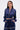 A woman wears the CALL BACK SATIN BOMBER, a shiny navy blue cropped satin jacket with pearl details and matching high-waisted pants. She has long straight hair, statement earrings, a serious look, and her hands in her pockets.