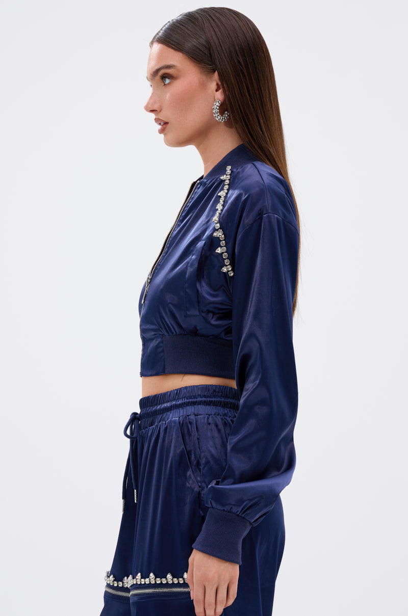 A woman with straight brown hair and statement earrings stands in profile, wearing the CALL BACK SATIN BOMBER—a shiny, navy blue cropped bomber jacket with rhinestone trim—paired with matching high-waisted pants.
