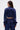 A woman with long brown hair wears the CALL BACK SATIN BOMBER in shiny navy blue with a cropped fit and matching high-waisted pants, facing away to display the back against a plain white background.