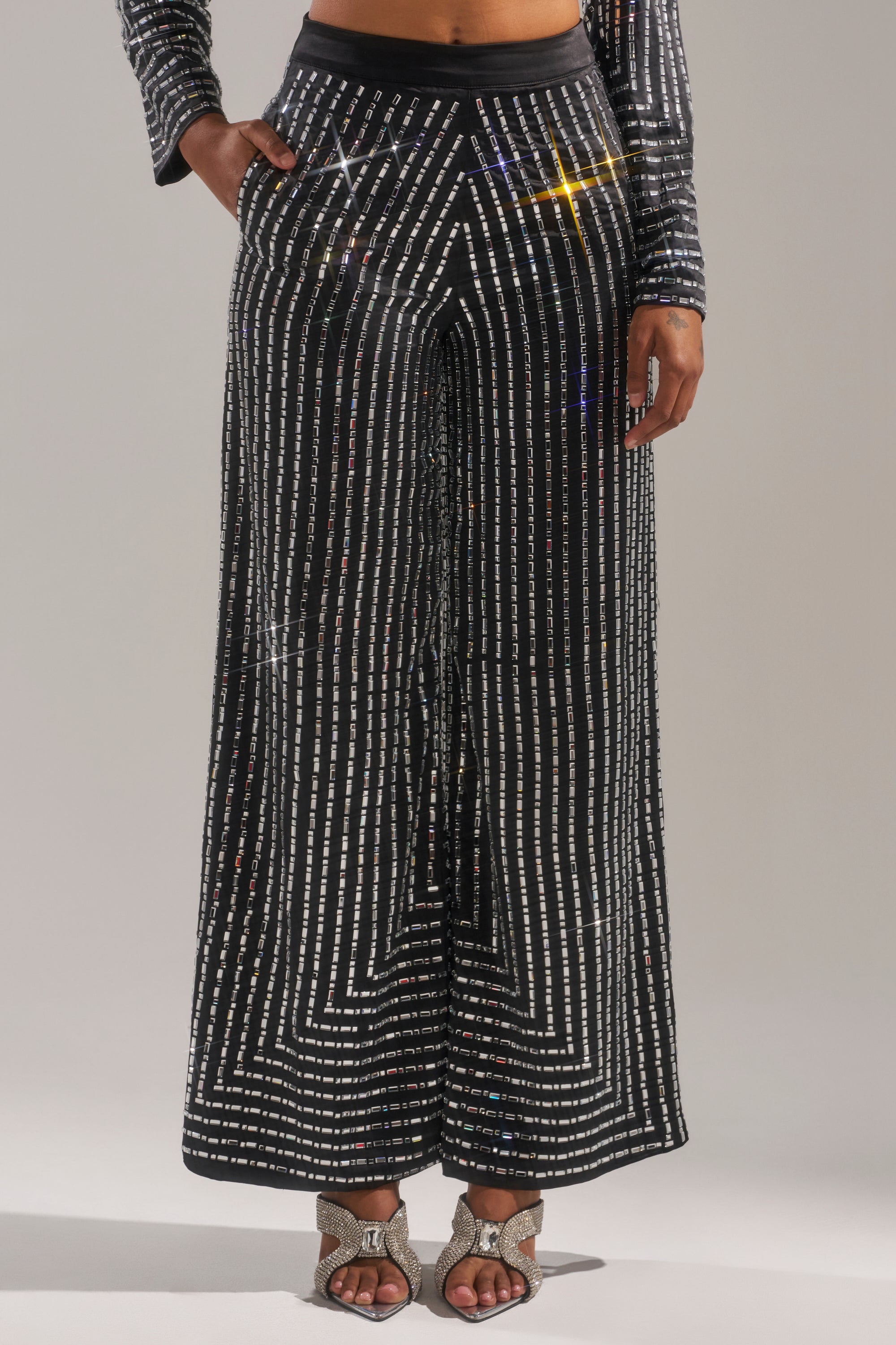 Someone wears the DONT STARE TOO HARD RHINESTONE TROUSER, a black high-rise wide-leg pant with silver sequin stripes, paired with a matching top and sparkly open-toe heels with bows, standing against a plain background.