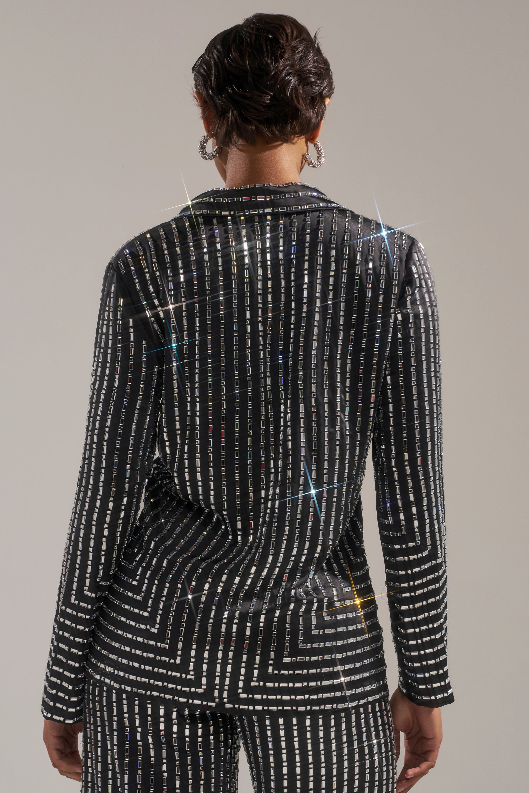 Seen from behind, a person with short hair wears the DONT STARE TOO HARD RHINESTONE BLOUSE, its black fabric covered in reflective rhinestones and geometric silver stripes, creating a dazzling effect against a plain background. 3