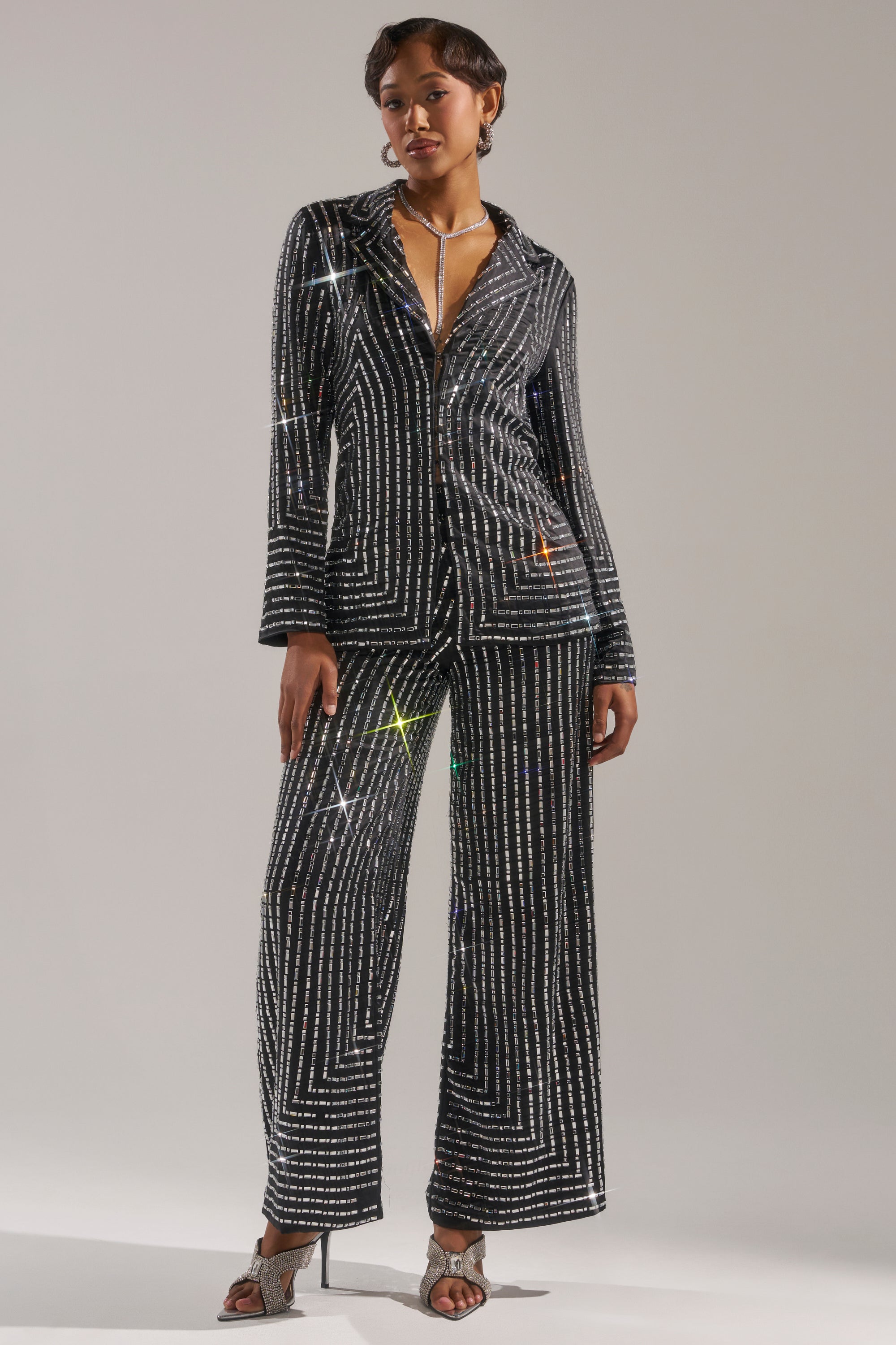 A woman exudes confidence in the DONT STARE TOO HARD RHINESTONE BLOUSE, featuring black fabric with sparkling geometric patterns, styled with wide-leg trousers, a silver necklace, and heeled sandals against a neutral backdrop. 4