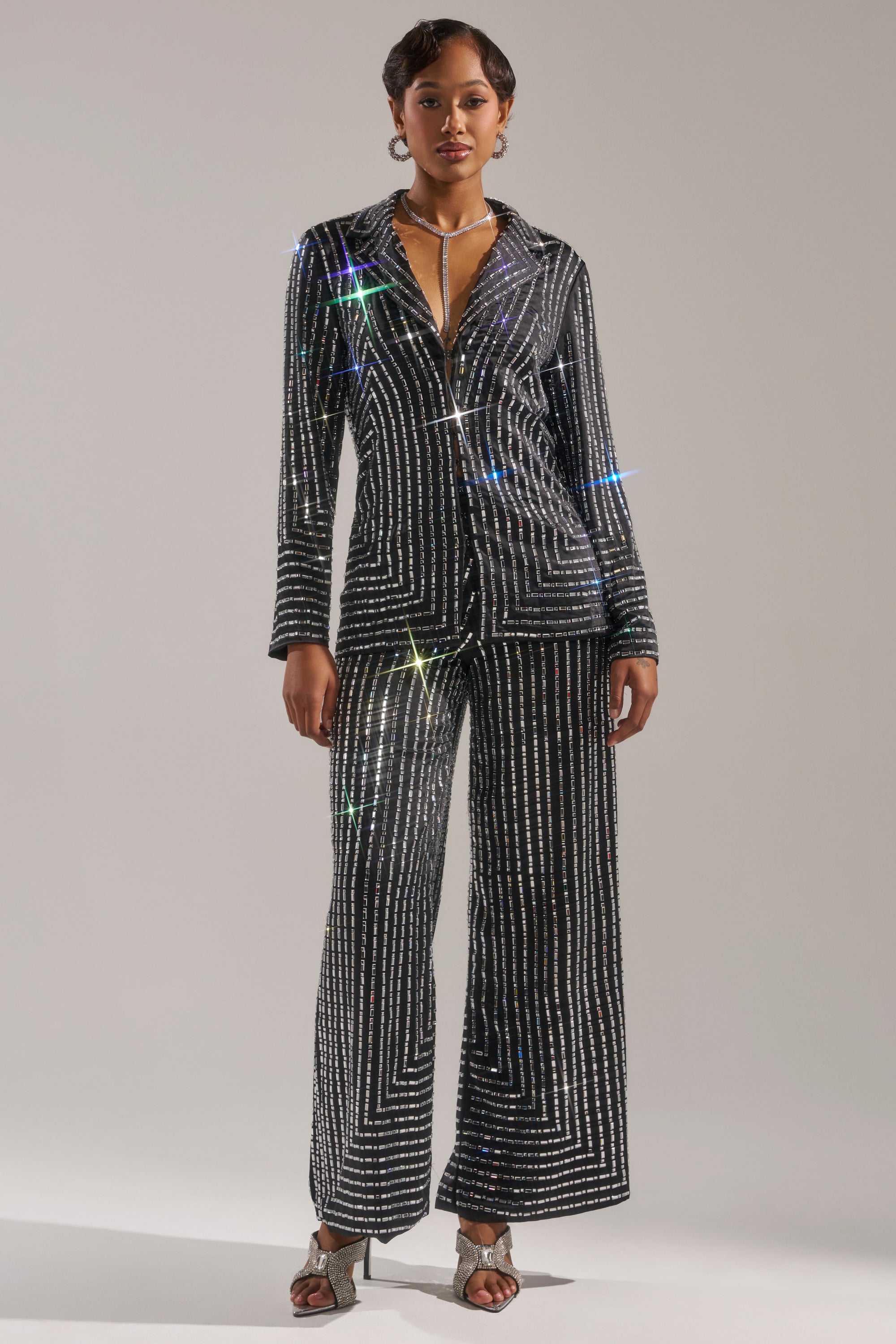 A woman wears the DONT STARE TOO HARD RHINESTONE BLOUSE with matching wide-leg pants, both covered in sparkling rhinestones. The black fabric catches the light, creating prism-like glimmers. She pairs the outfit with silver heels and a necklace. 5
