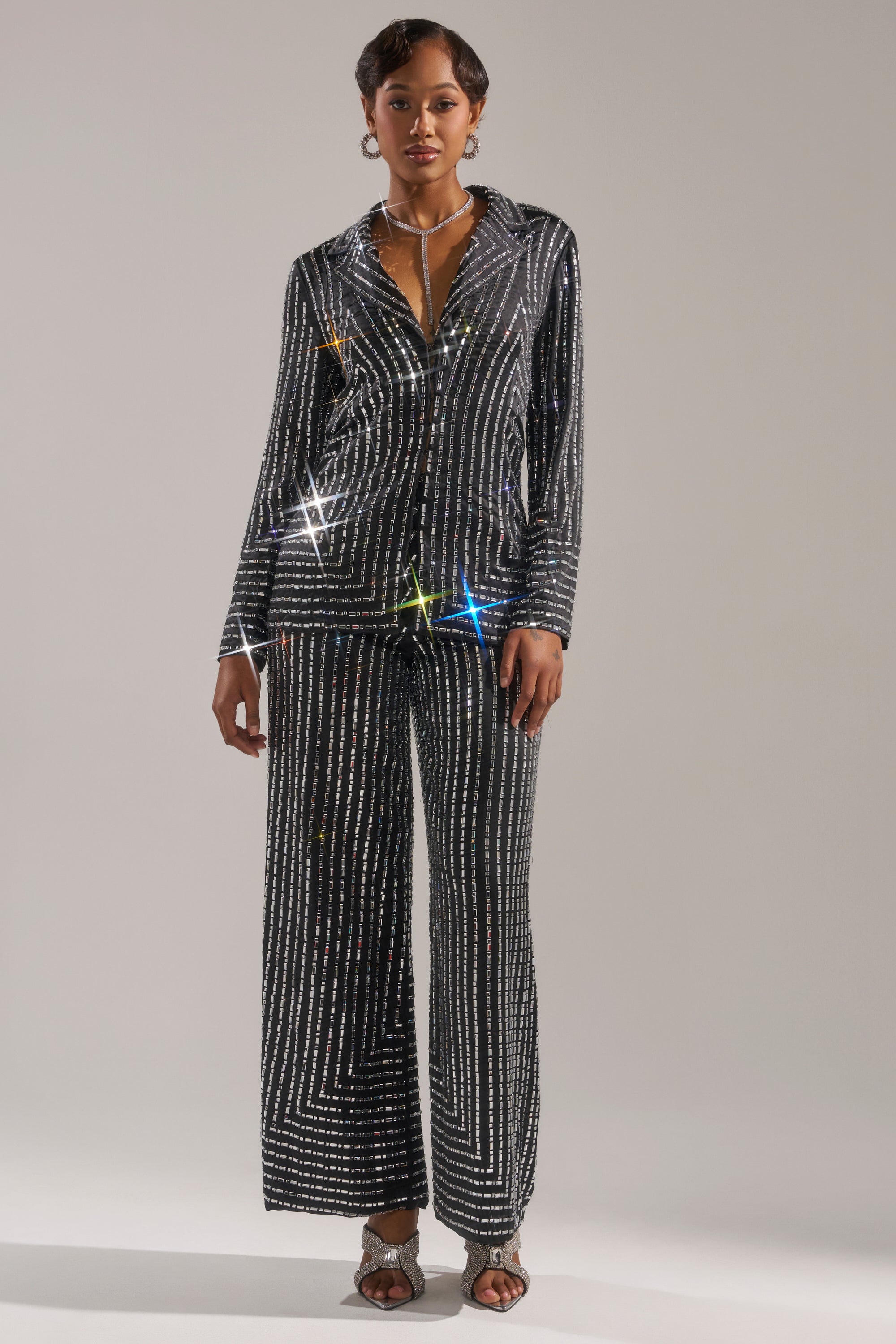 A woman with short hair and minimal jewelry poses against a light gray backdrop, modeling the AKIRA Label DONT STARE TOO HARD RHINESTONE BLOUSE—an eye-catching black, geometric-patterned top paired with wide-leg pants and silver heeled sandals. 6