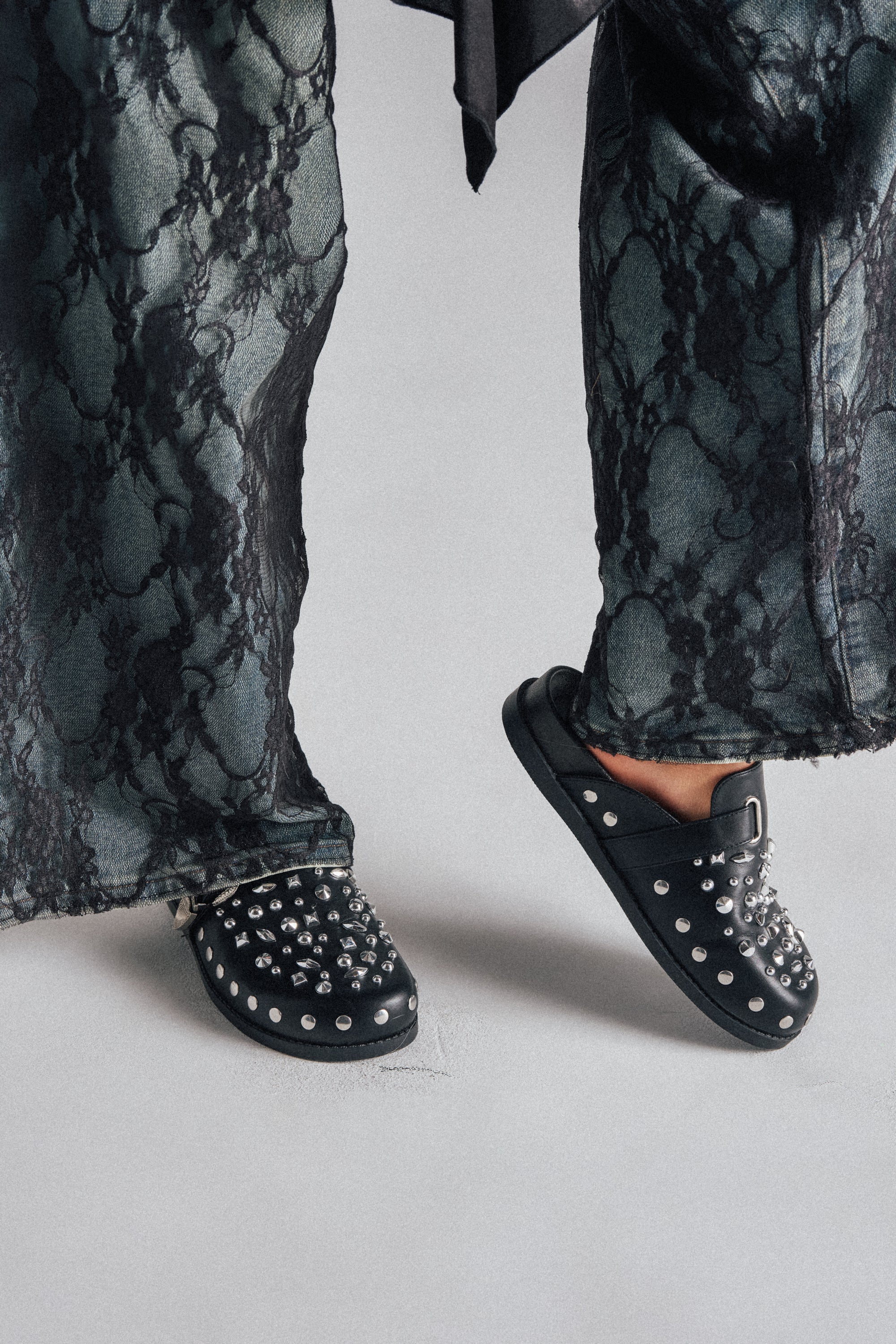 A person stands on a light gray surface wearing loose, dark patterned pants and the AZALEA WANG TURIN BLACK METAL EMBELLISHMENT FLAT shoes.
