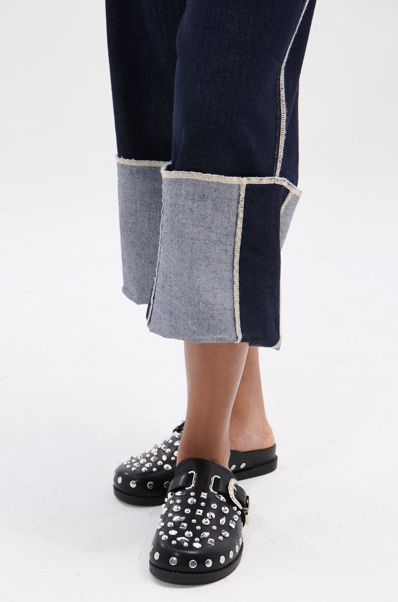 A person wears cropped, wide-leg jeans with exposed seams and two-tone panels, paired with the AZALEA WANG TURIN BLACK METAL EMBELLISHMENT FLAT slip-ons featuring a western-style buckle and metallic studs.