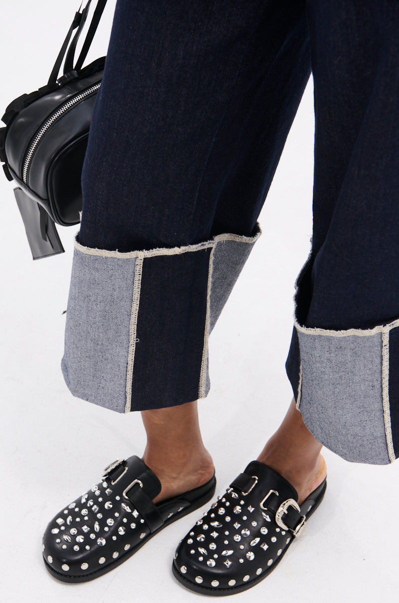 A person stands on a white surface wearing wide-leg cuffed dark denim jeans and AZALEA WANG TURIN BLACK METAL EMBELLISHMENT FLAT shoes, holding a small faux leather black bag.