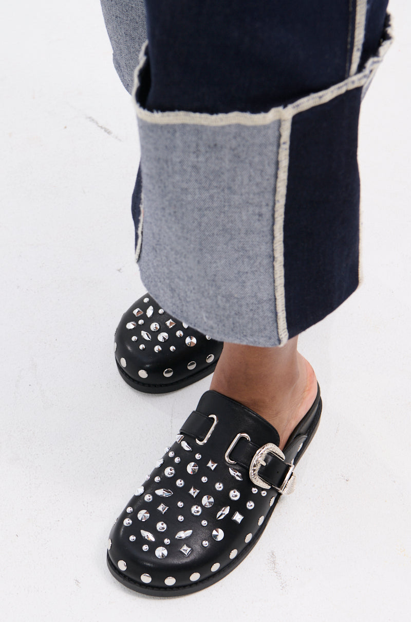 A person stands on a white surface wearing two-tone denim pants with large cuffs and black AZALEA WANG TURIN METAL EMBELLISHMENT FLATS featuring metallic studs and a rhinestone western-style buckle.