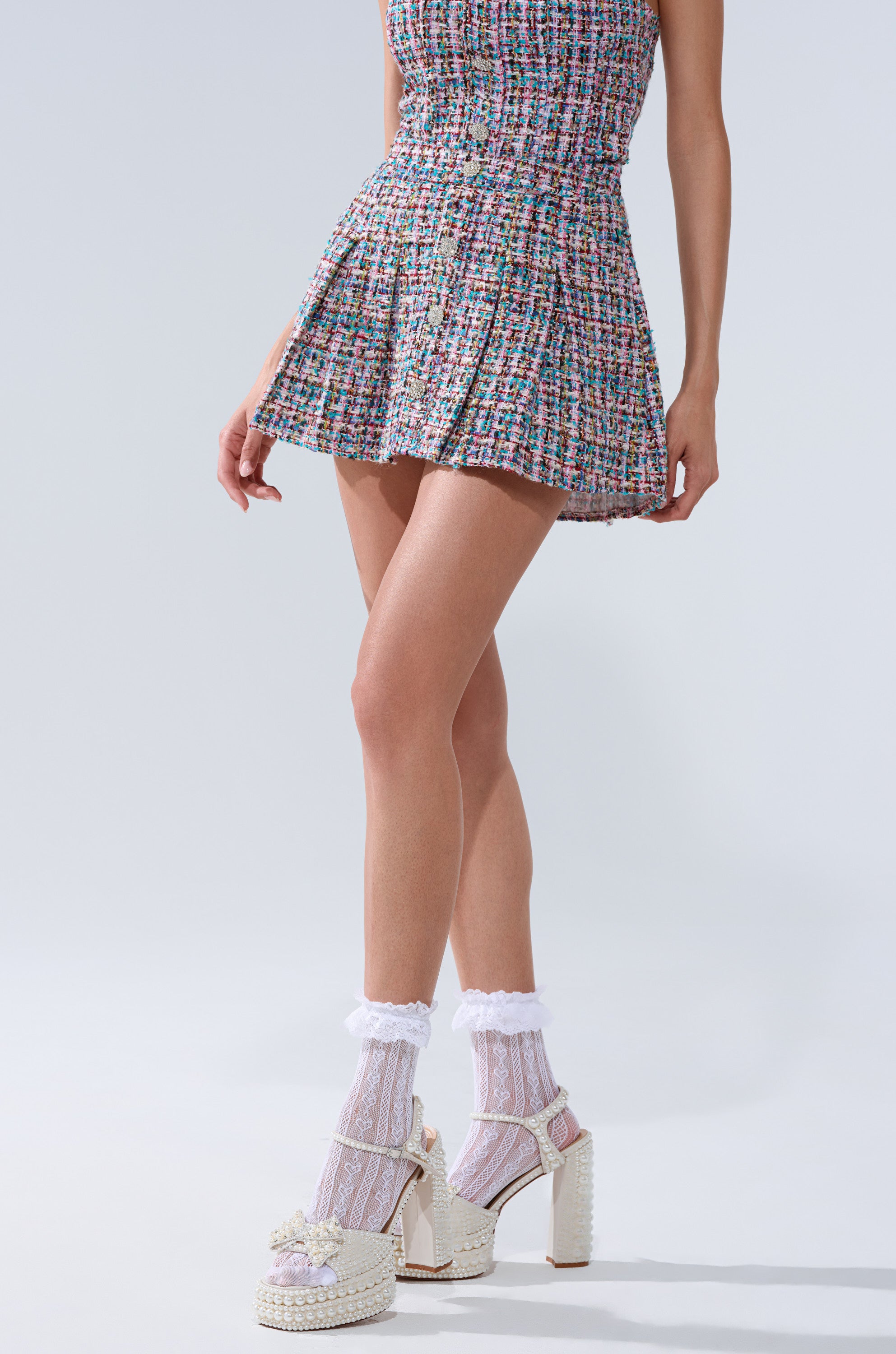 A woman wears the Tweed Pleated Mini Skirt with metallic thread, ruffled white lace socks, and pearl-embellished platform sandals with bows, standing against a plain light background. Only her lower half is visible.