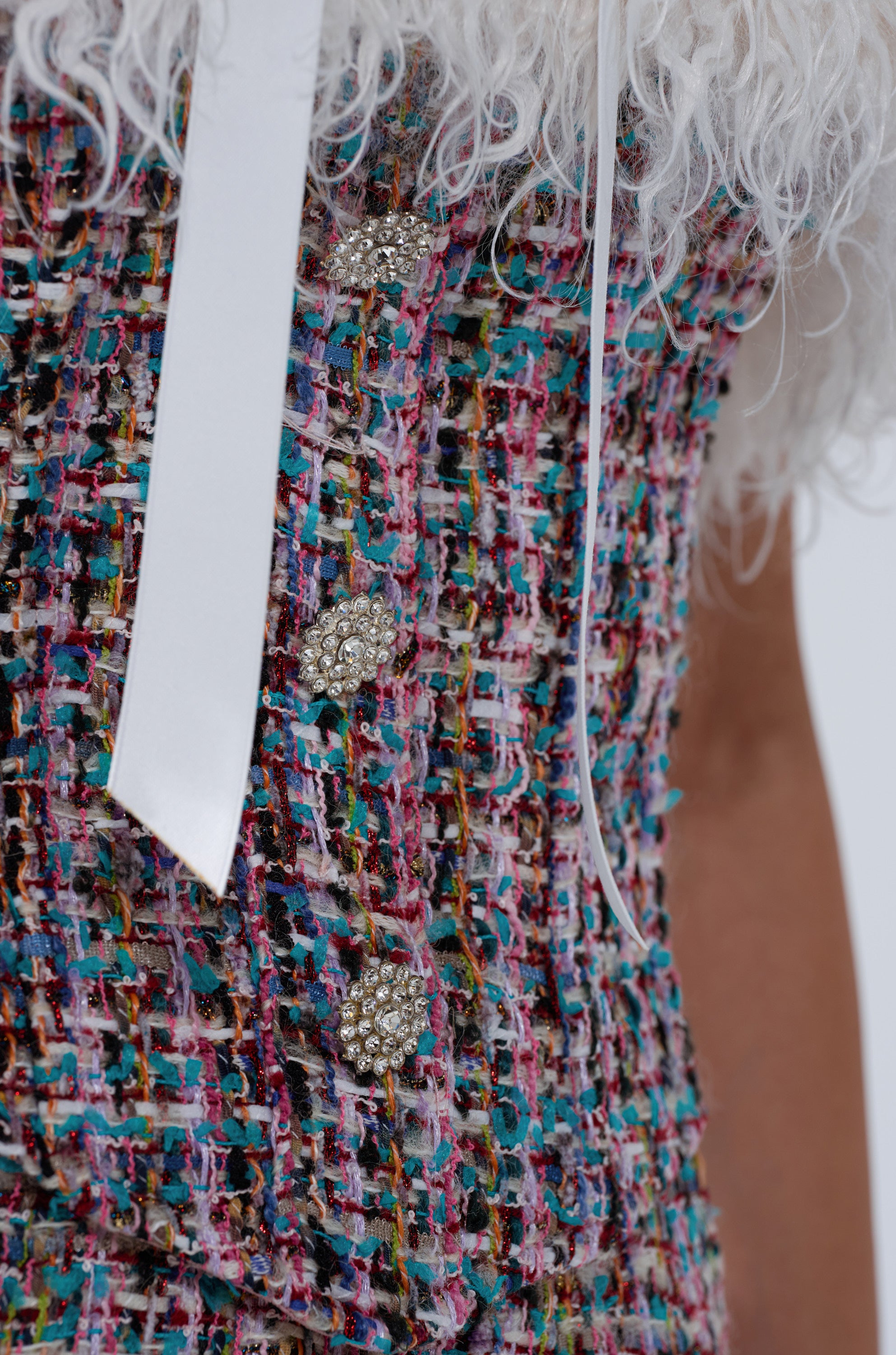 Close-up of the FUR TRIM TWEED CORSET featuring textured multicolored fabric, jeweled buttons, and a white ribbon, styled over a fluffy faux fur top. The image shows part of the wearer's torso and arm.