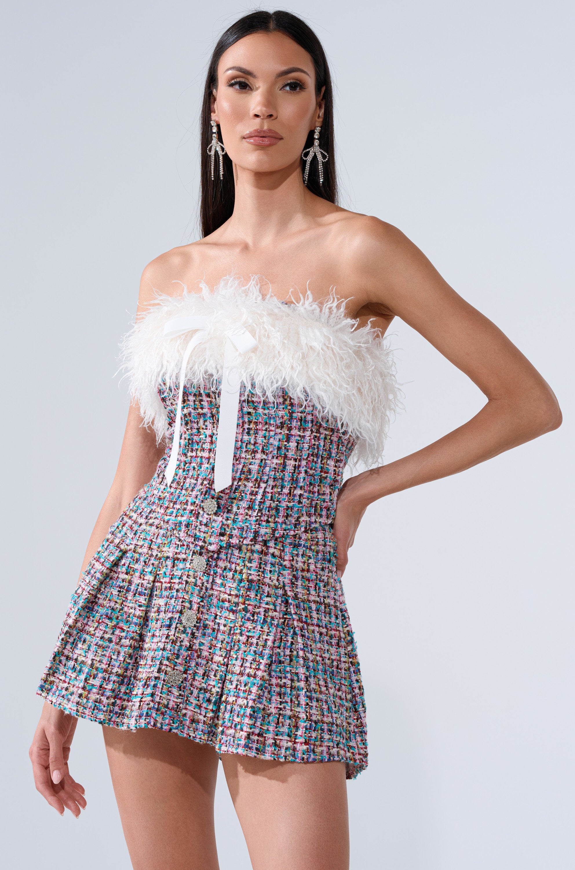 A woman models the FUR TRIM TWEED CORSET, a strapless mini dress with a vibrant tweed pattern, feathered faux fur at the neckline, and a white ribbon accent. She has long straight hair and bold statement earrings.